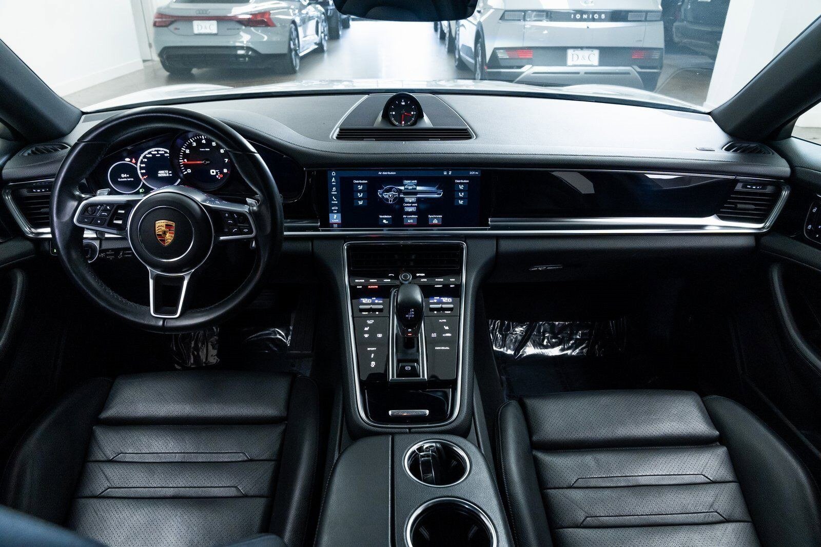 Interior of a luxury car cockpit showing a leather-wrapped steering wheel with Porsche emblem, digital instrument cluster, central touchscreen display, gear shifter, and climate control buttons, with parked cars visible through the windshield.