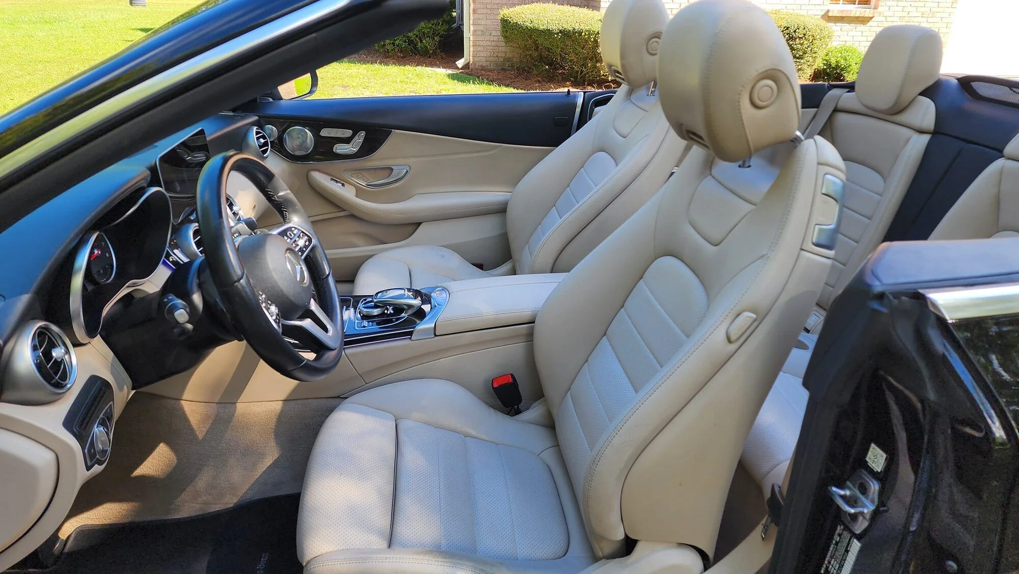 Interior of a luxury convertible car with beige leather seats and a black dashboard, parked outdoors with houses and greenery visible in the background.