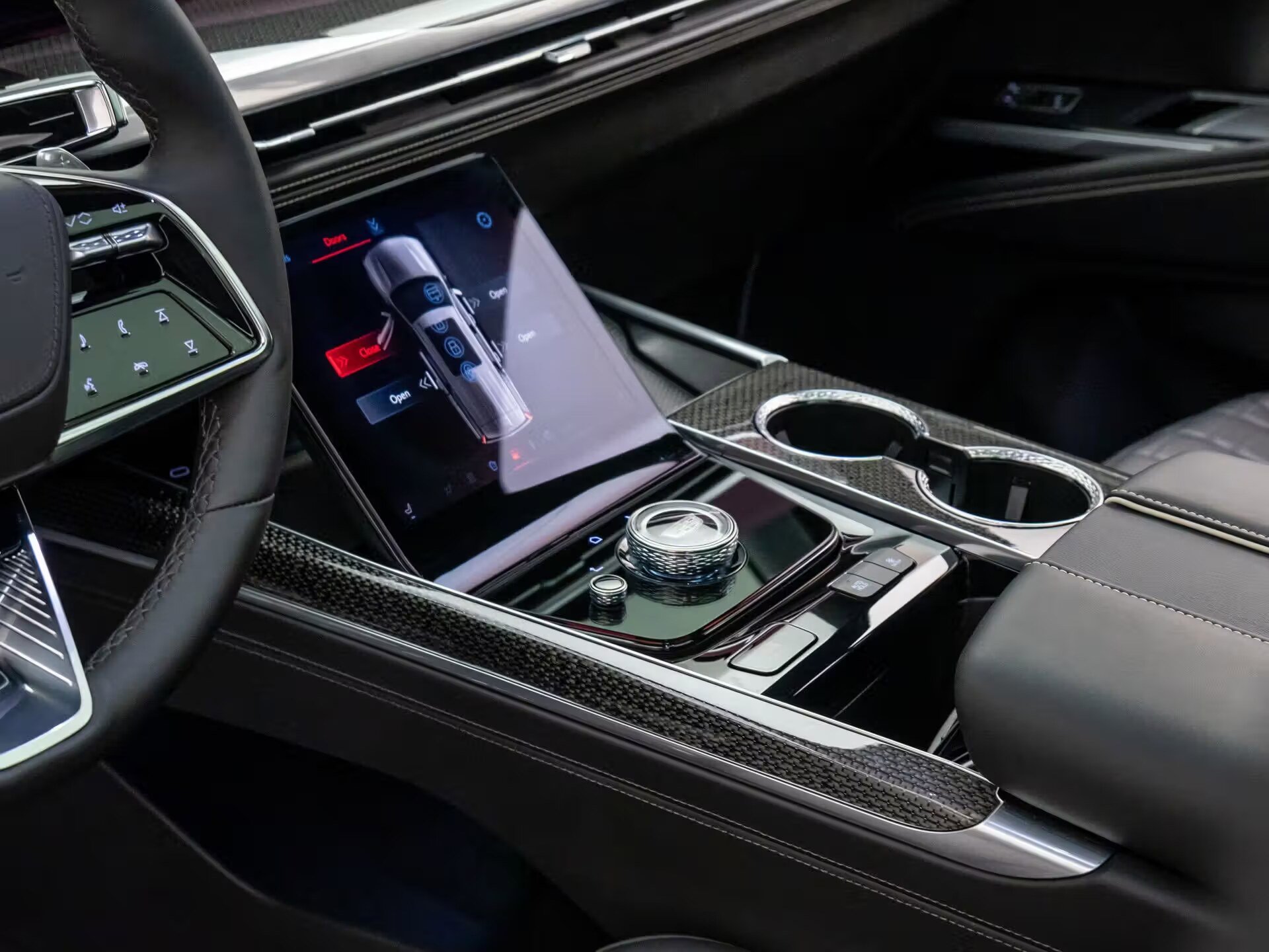 Interior of a luxury car with a digital touchscreen display, a rotary gear shifter, and cup holders, featuring black and silver accents.