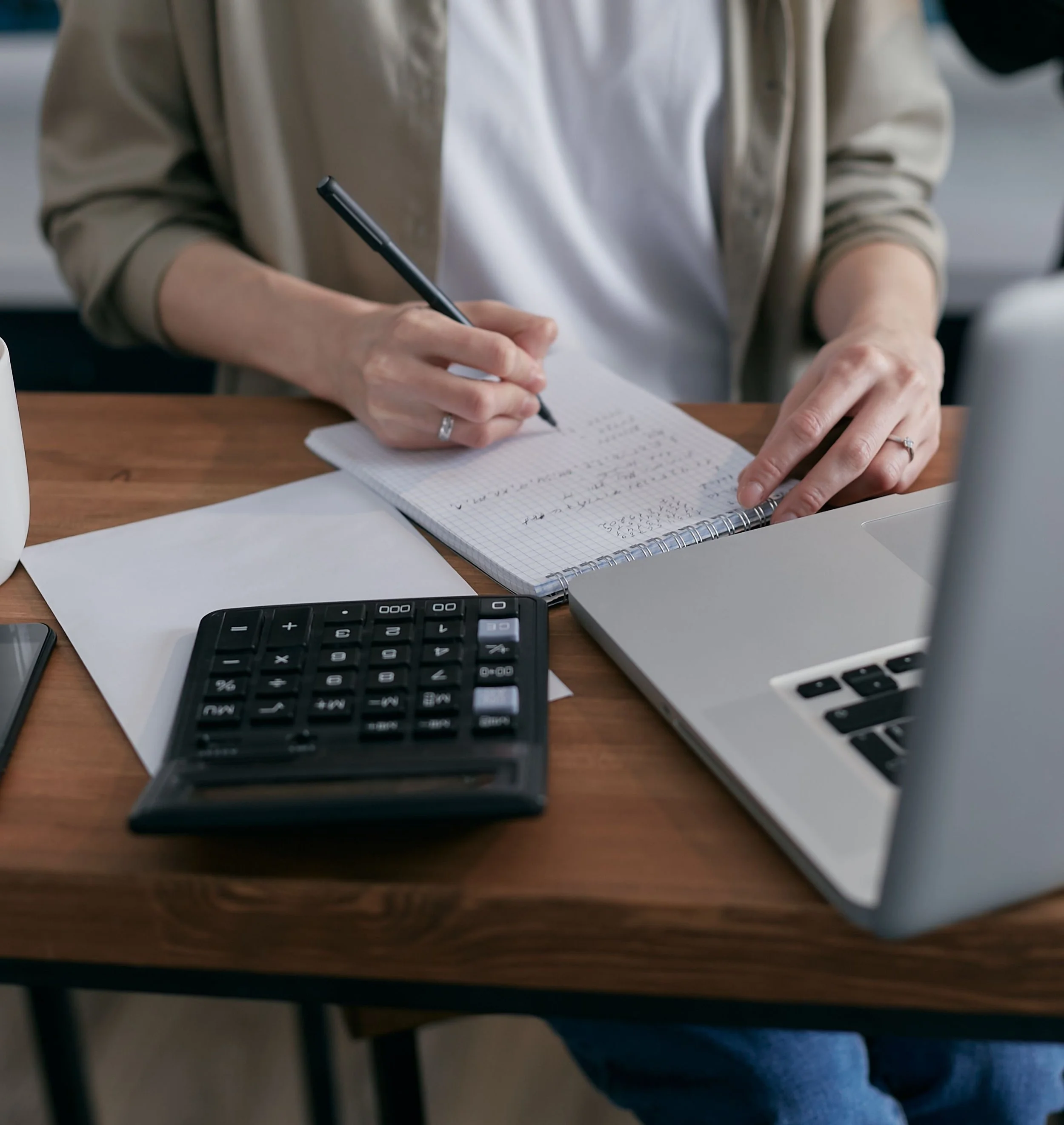 Close-up of a person writing in a spiral notebook at a wooden desk, with a calculator and a laptop nearby, in an indoor setting.