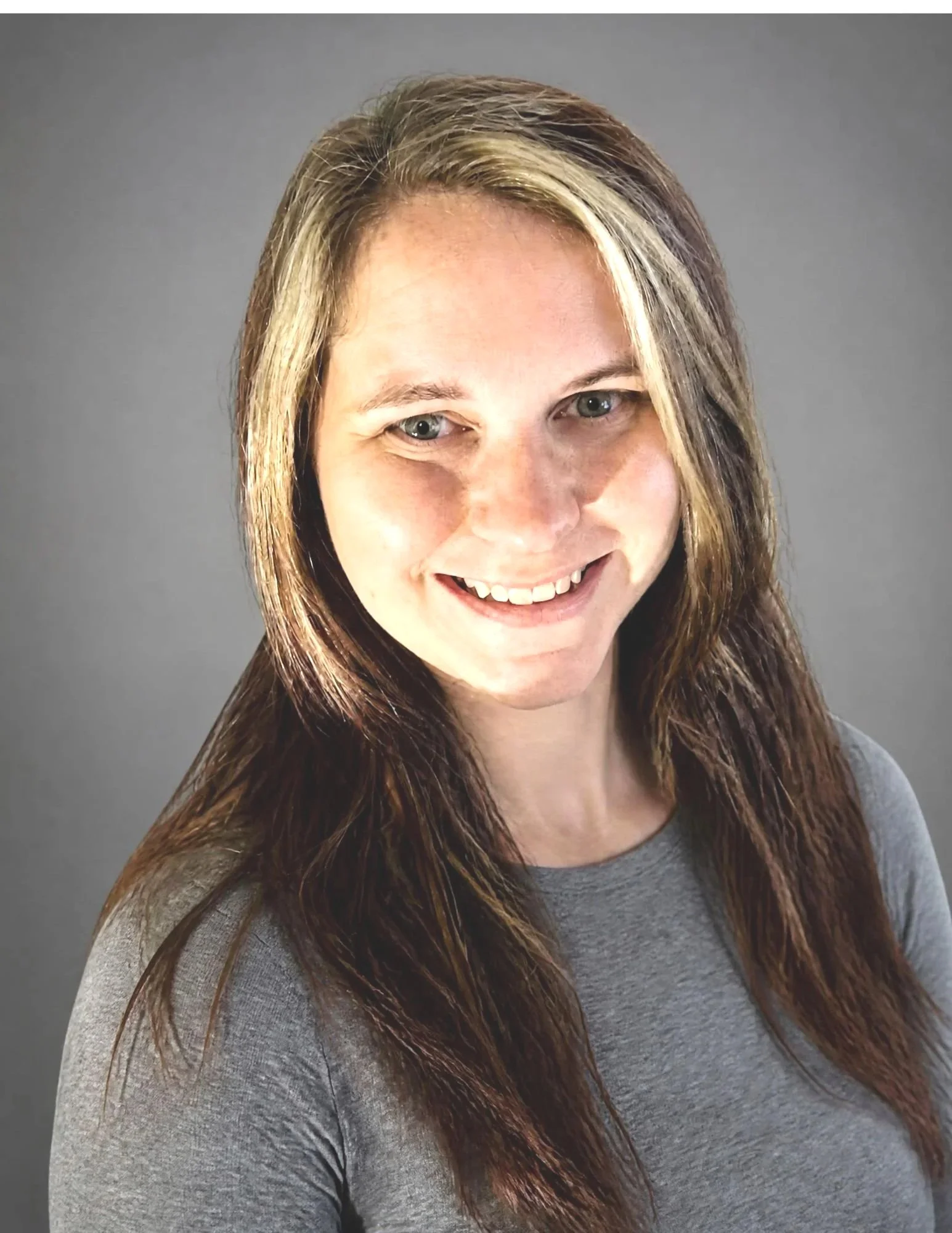 A woman with long, brown hair and blue eyes smiling, wearing a gray shirt, against a plain gray background.