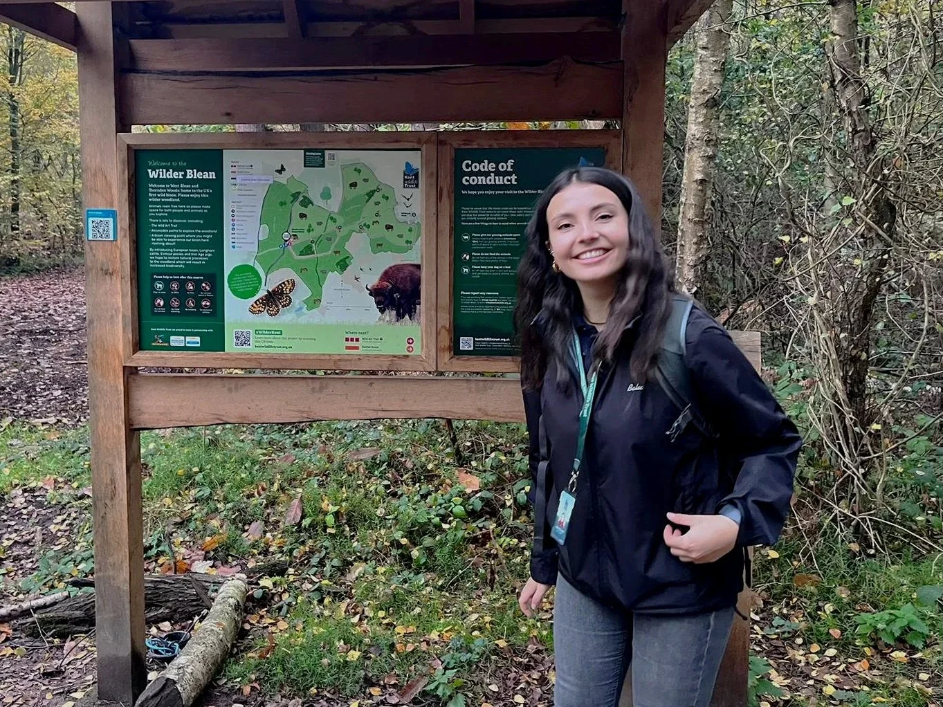 A woman with long dark hair smiling at the camera in front of a wooden signboard in a wooded park area. The signboard displays a map and information about the Wilder Blean nature reserve, including a code of conduct and a butterfly illustration.