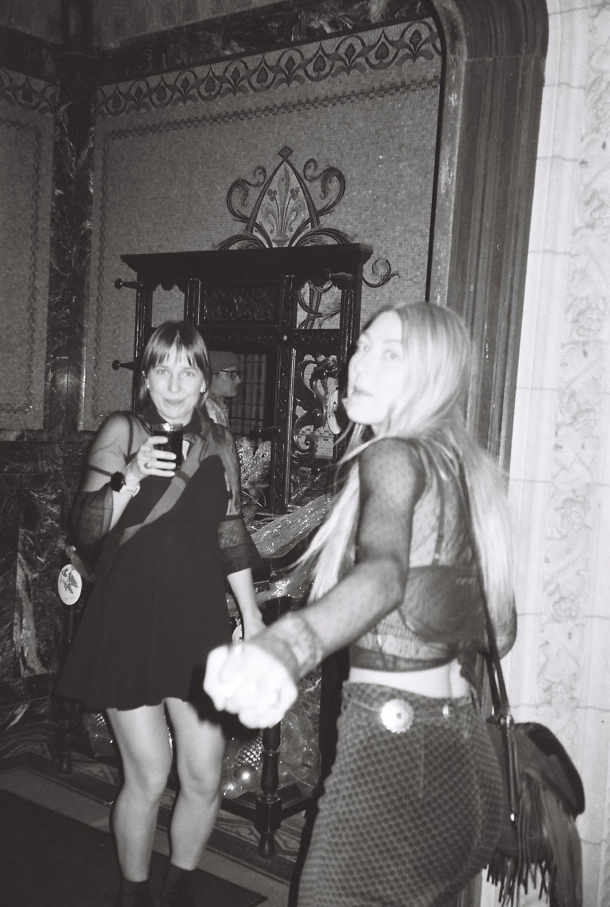 Two women holding hands and dancing at a social gathering or party indoors, with one woman holding a drink and smiling at the camera, and the other woman facing sideways with long hair and a patterned dress.
