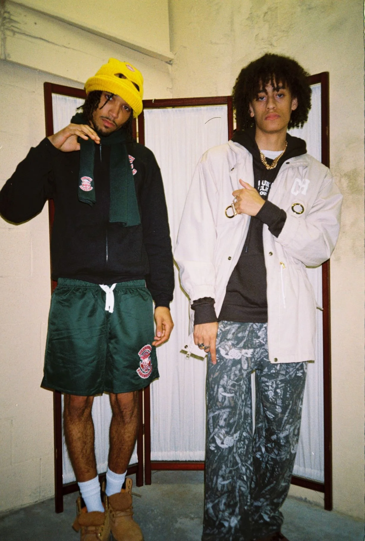Two young men standing indoors, posing in front of a folding room divider. The man on the left is wearing a yellow beanie, black jacket, green shorts, and tan sneakers with white socks. The man on the right has curly hair, is wearing a white jacket o