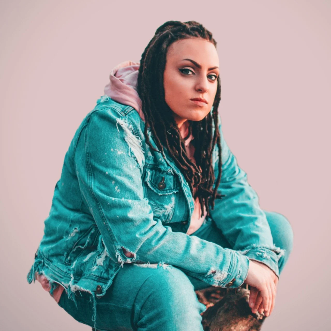A young woman with long dreadlocks wearing a distressed denim jacket and pants, sitting against a plain background.