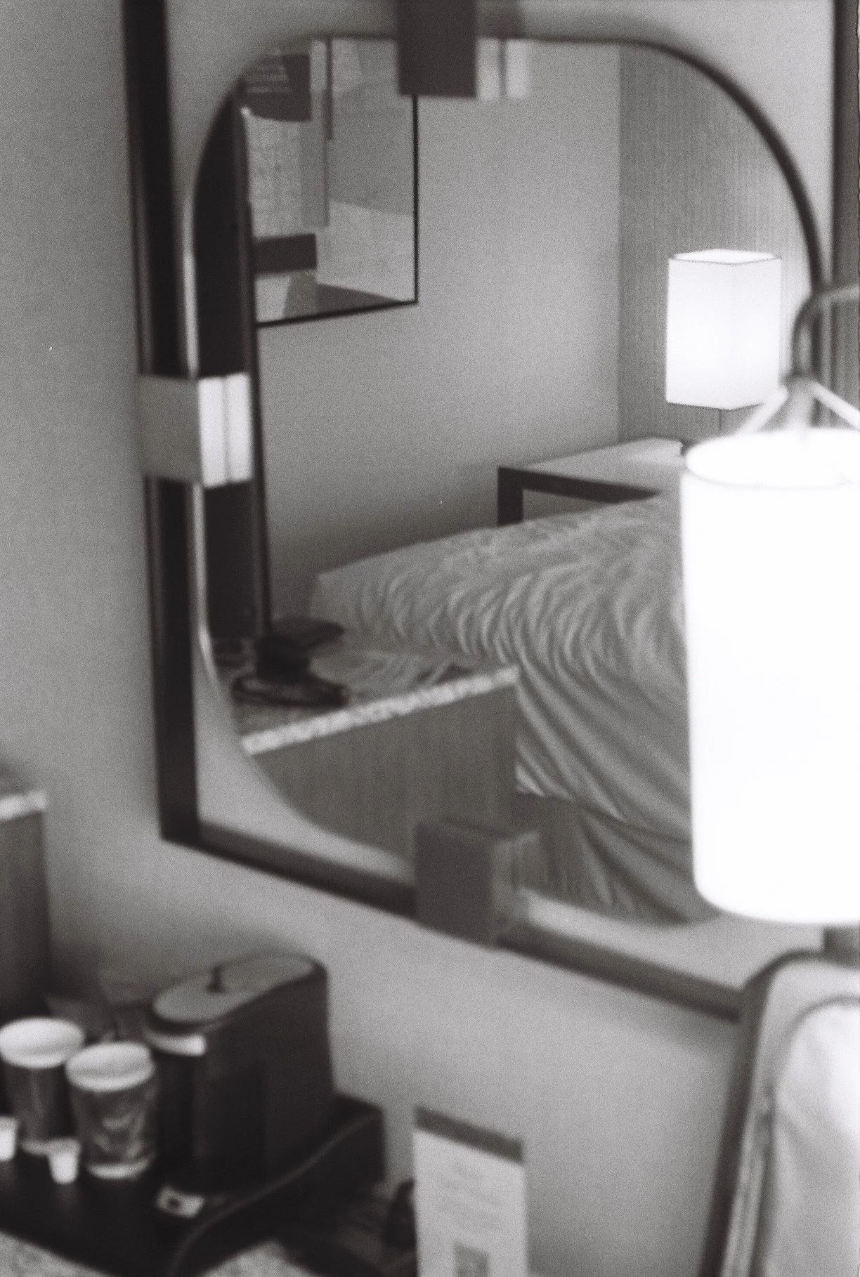 A hotel room as seen through a mirror reflecting a bed with a striped blanket, a nightstand with a lamp, and a framed picture on the wall.
