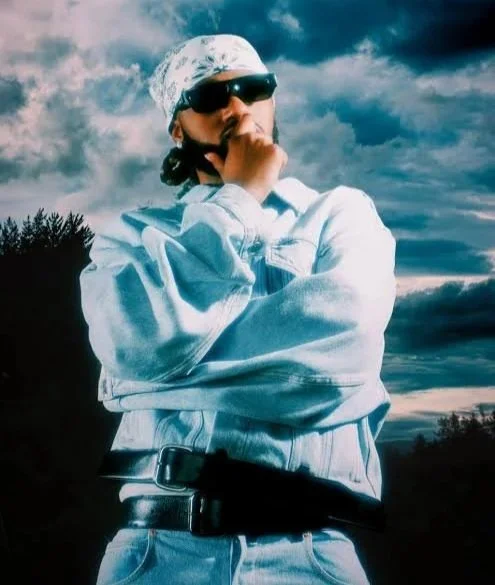 Person wearing sunglasses and a bandana, standing outdoors with a cloudy sky background.