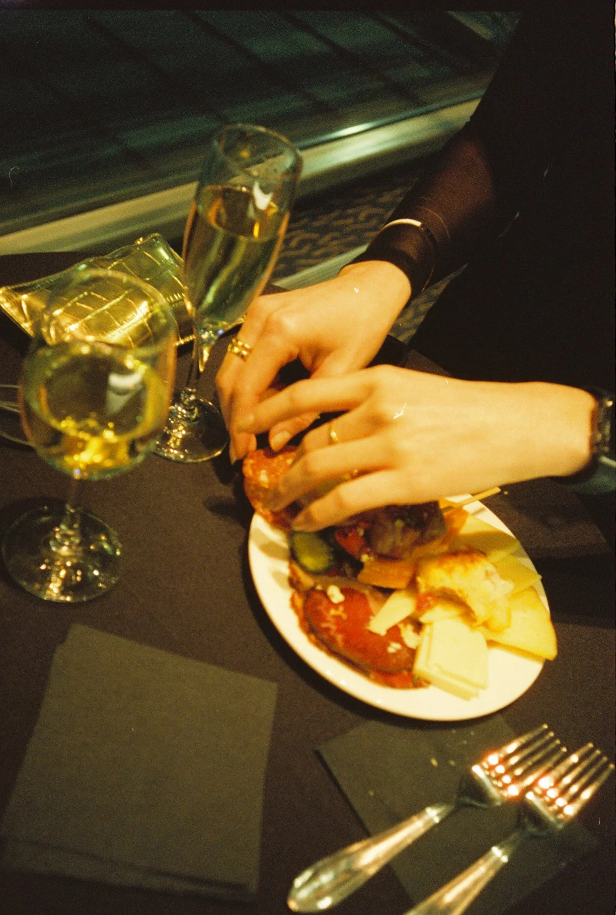 A person eating a plate of assorted appetizers or finger foods at a dark table with two glasses of champagne nearby.