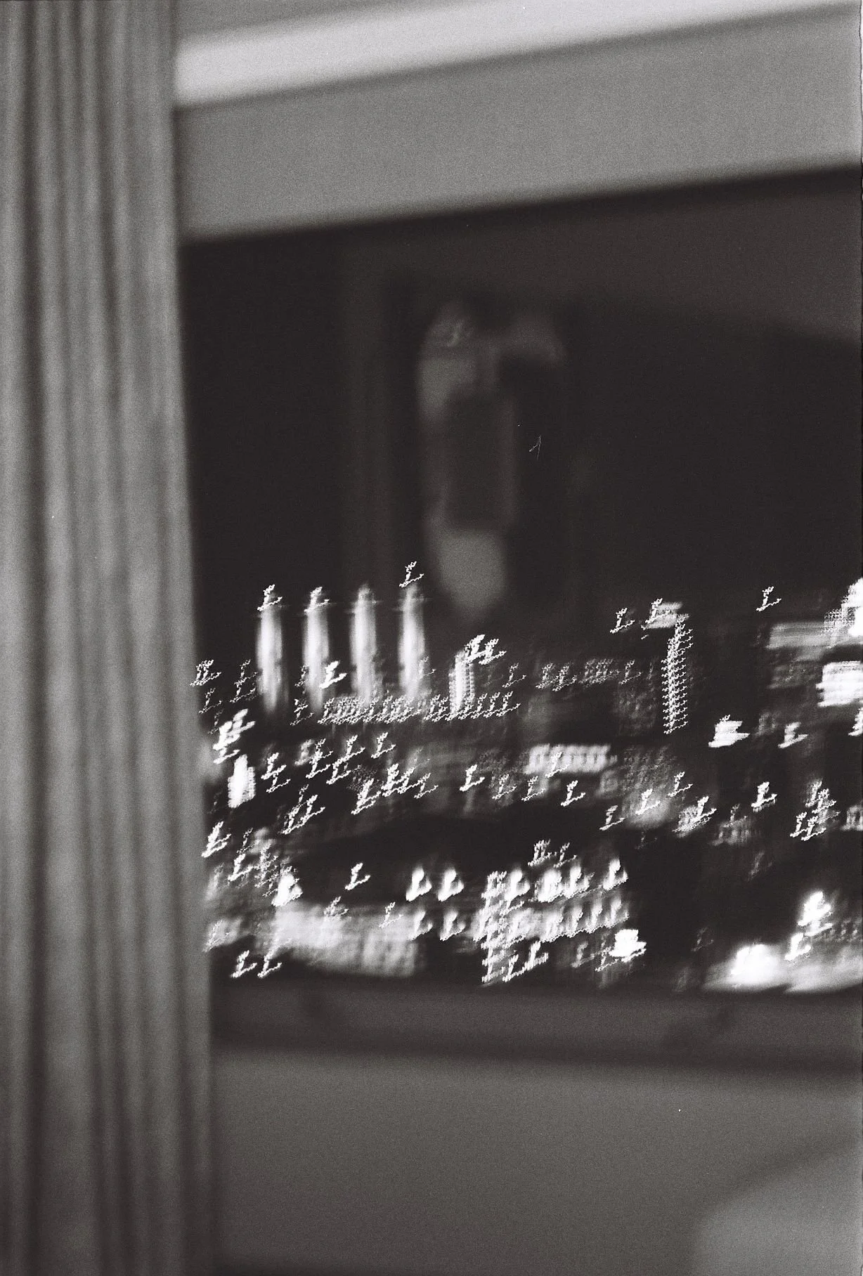 Black and white photo of city lights seen through a window at night with a reflection of a person taking the photo.