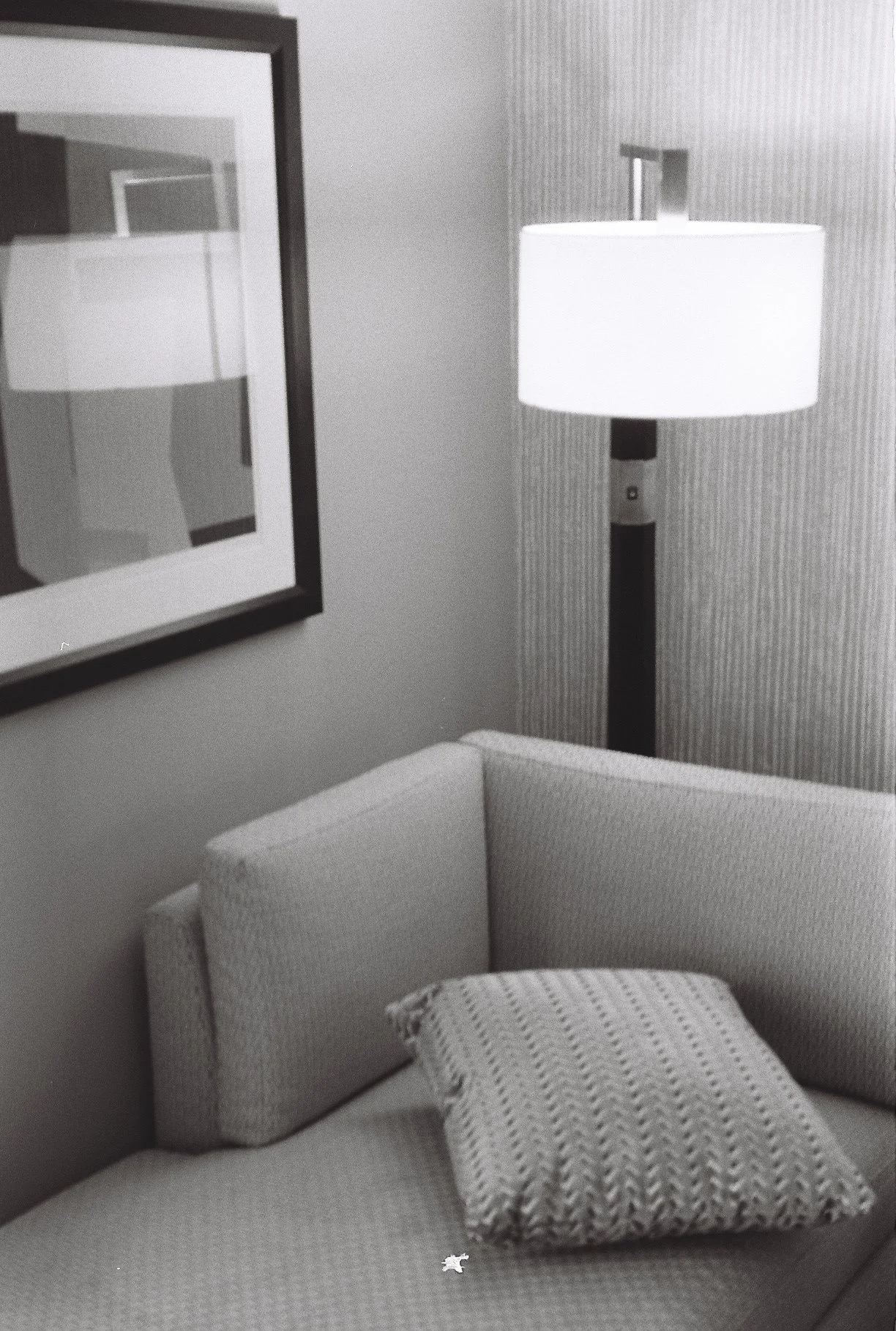 A living room with a beige upholstered sofa, a patterned cushion, a floor lamp with a white shade, a framed abstract artwork on the wall, and a textured wallpaper.