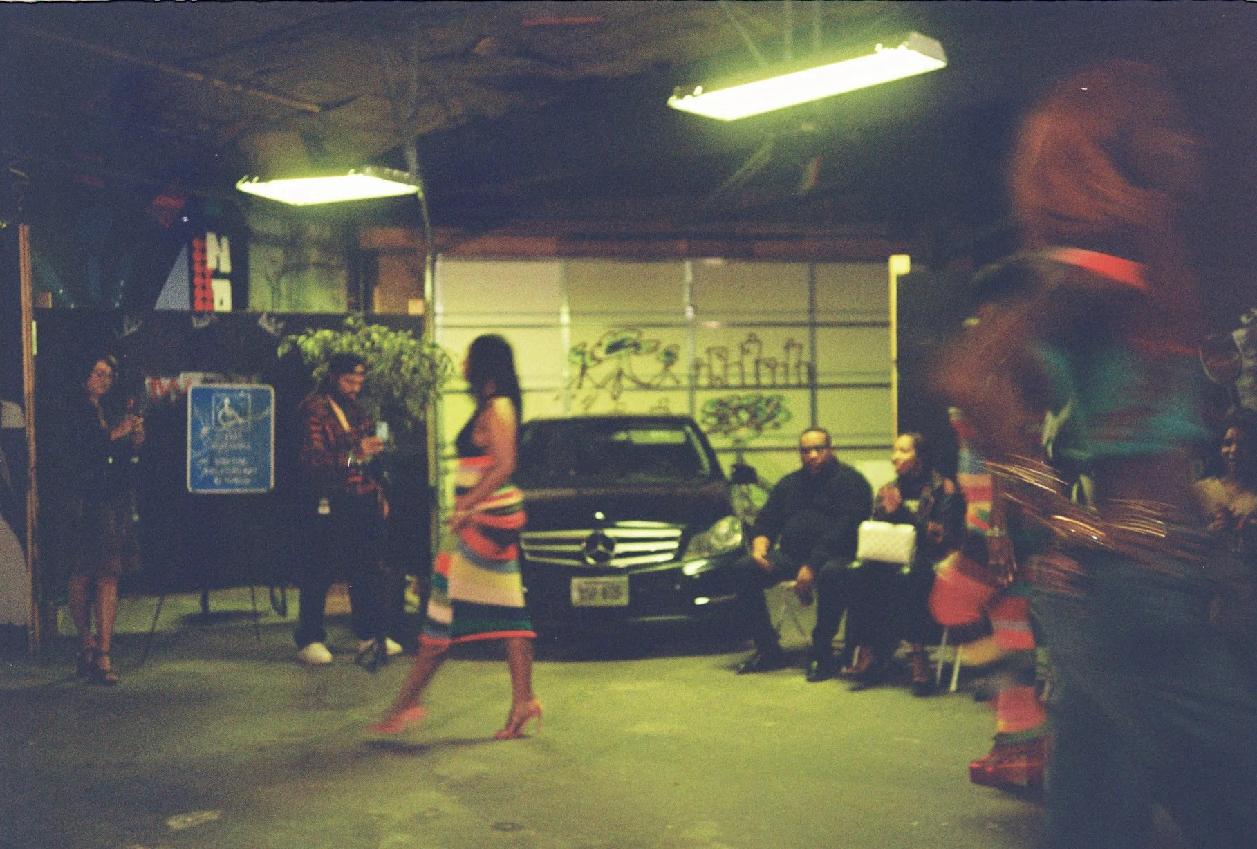 Nighttime scene with multiple people standing and sitting outside a garage, some holding drinks. A black Mercedes-Benz is parked in front of the garage which has shelves with bottles on the wall. People are dressed casually, and a woman in a striped 