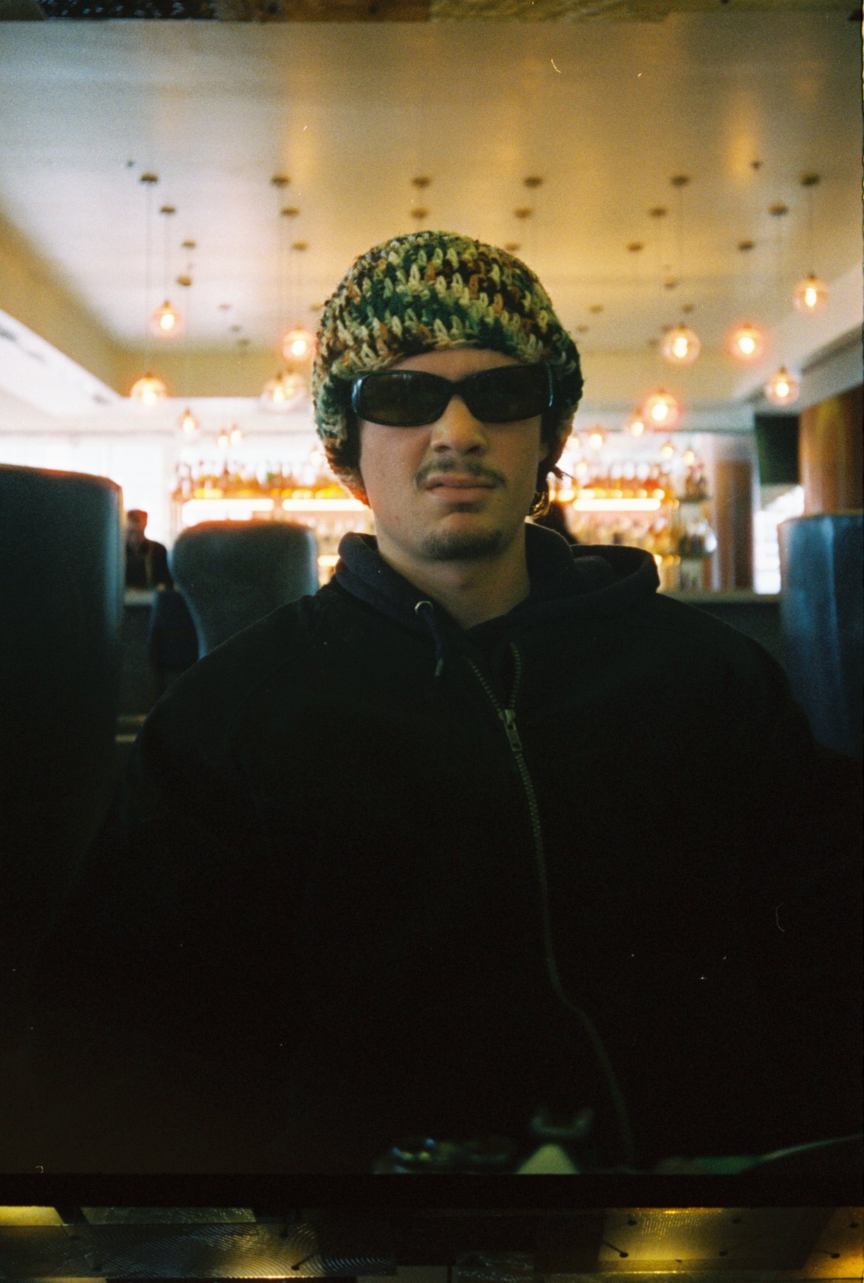 Man wearing sunglasses and a patterned hat, sitting in a dimly lit restaurant or bar with warm lighting, behind a computer monitor.