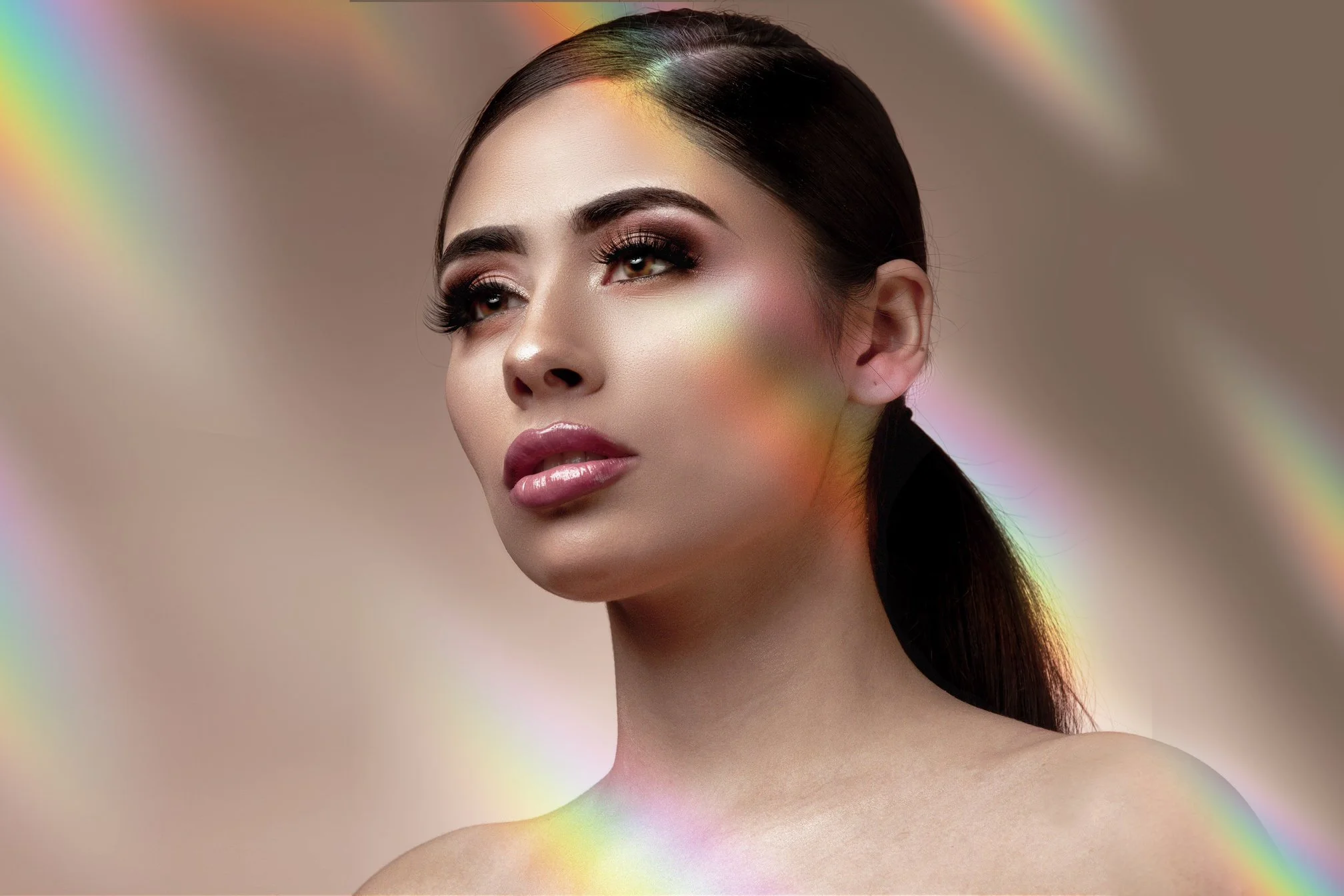 A close-up portrait of a woman with dark hair styled in a sleek ponytail, wearing makeup with dark eyeshadow, long lashes, and glossy lips, with colorful light reflections on her face and background.