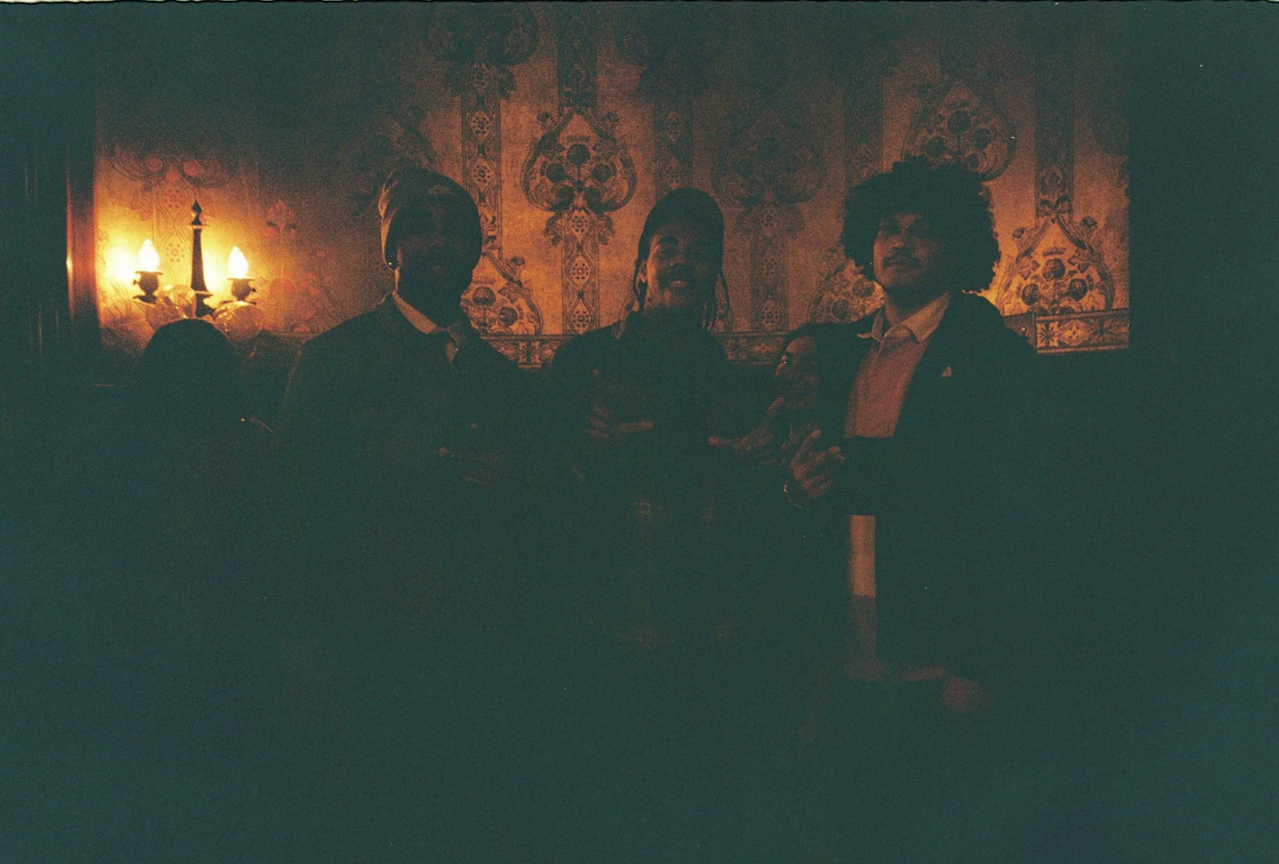 Three people standing together in a dimly lit room with ornate wallpaper and a wall sconce with two lamps.
