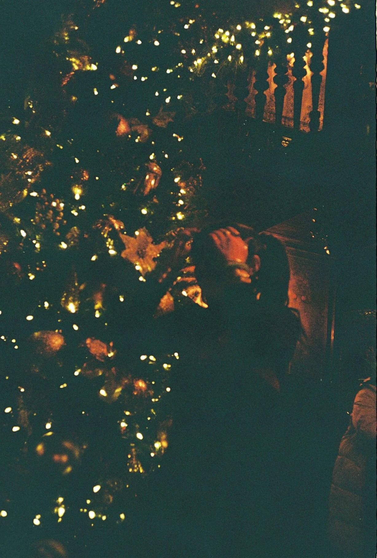 A person arranging Christmas decorations on a Christmas tree at night, with warm lighted ornaments. There is a wooden railing in the background.