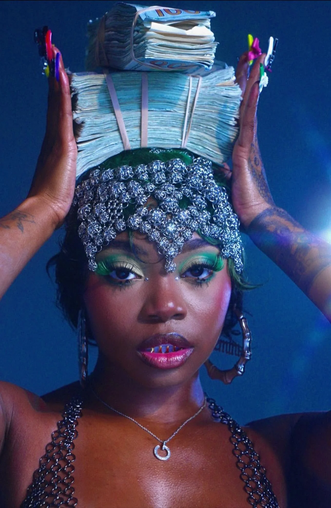 A woman with elaborate makeup and jewelry holding a stack of money balanced on her head. She has distinctive colorful eyeshadow, visible tattoos, and neon-colored nails.