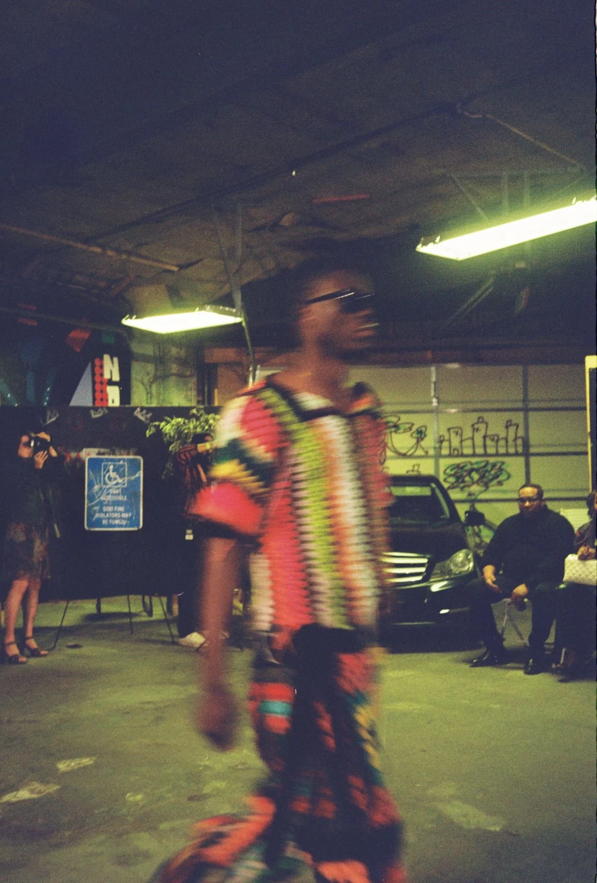 A woman in colorful clothing walking through a parking garage with several seated people, a car, and graffiti on the wall.