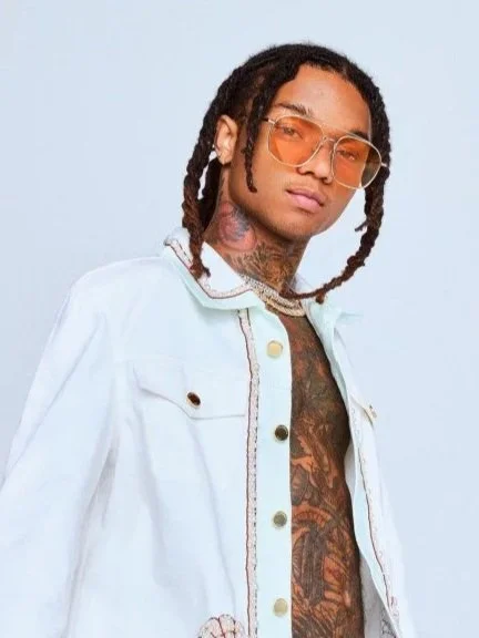 A young man with dreadlocks, wearing tinted glasses and a white denim jacket, looking at the camera against a light blue background.