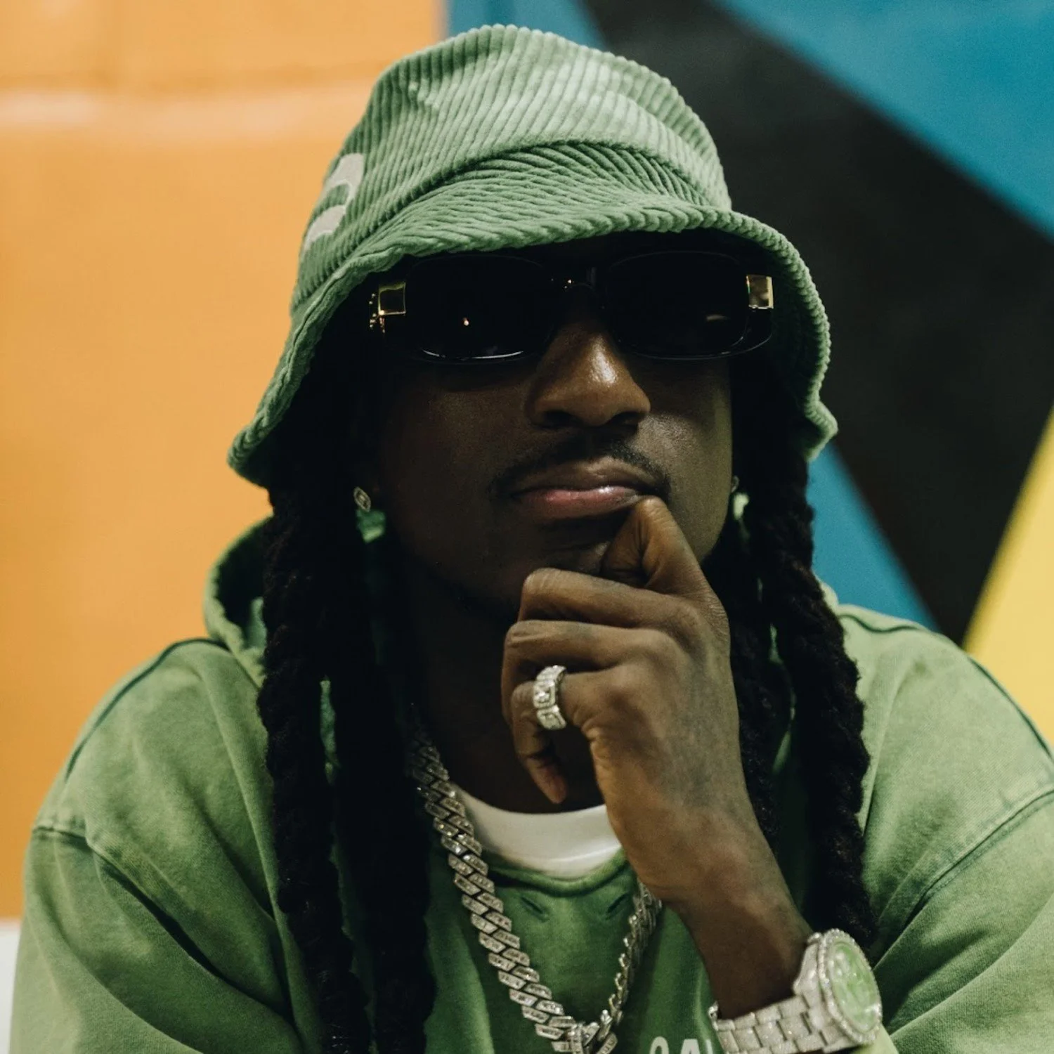 A man wearing a green bucket hat, black sunglasses, and a green sweatshirt, with long dreadlocks, jewelry, and a watch, resting his chin on his hand in front of a colorful background.