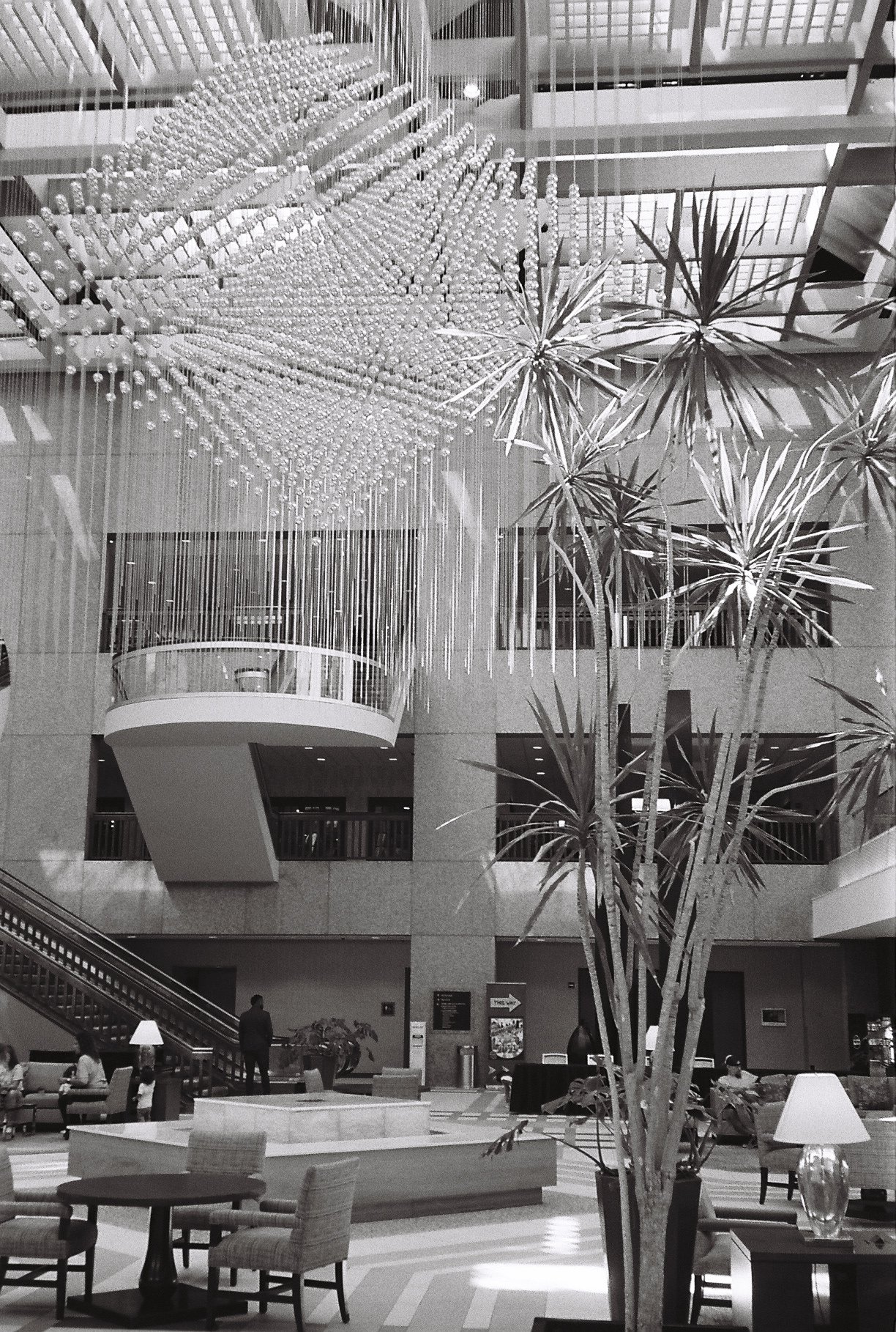 Interior of a hotel lobby with a large chandelier, potted plants, seating area, and people sitting and walking.