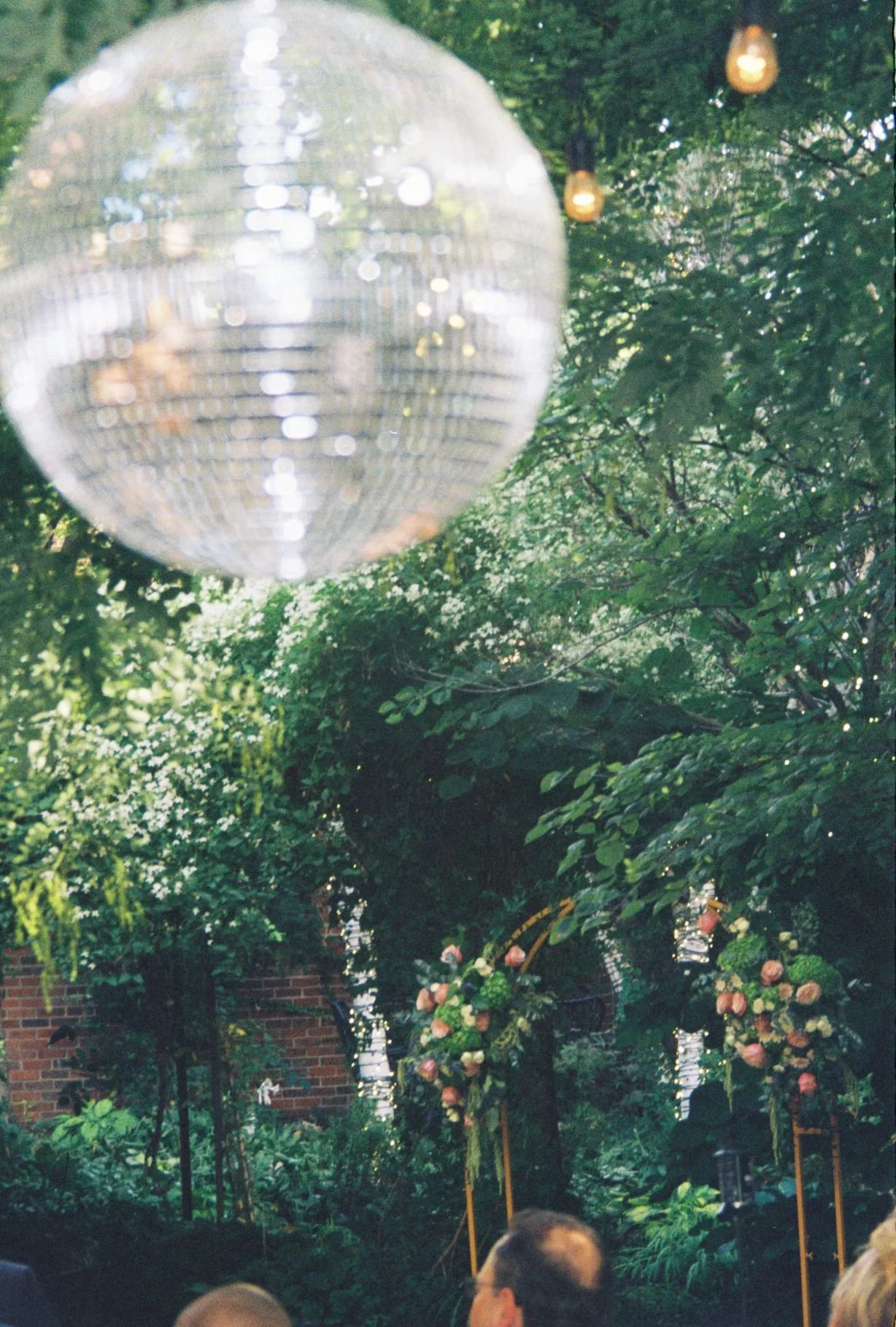 A large disco ball hanging from tree branches amidst string lights and greenery, with a garden setting and floral arrangements, likely at an outdoor event or celebration.