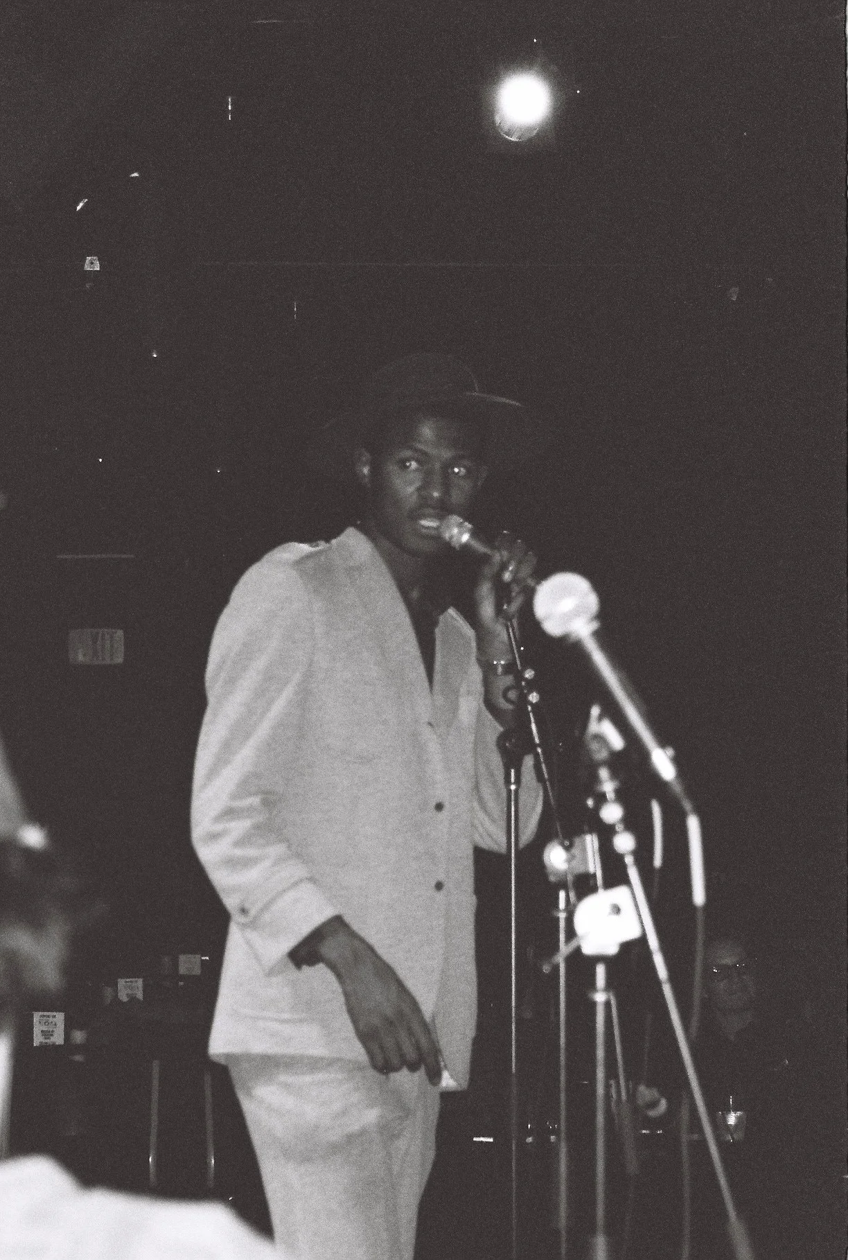 A person wearing a hat and light-colored clothing standing at a microphone in a dark setting, possibly a stage or performance space.