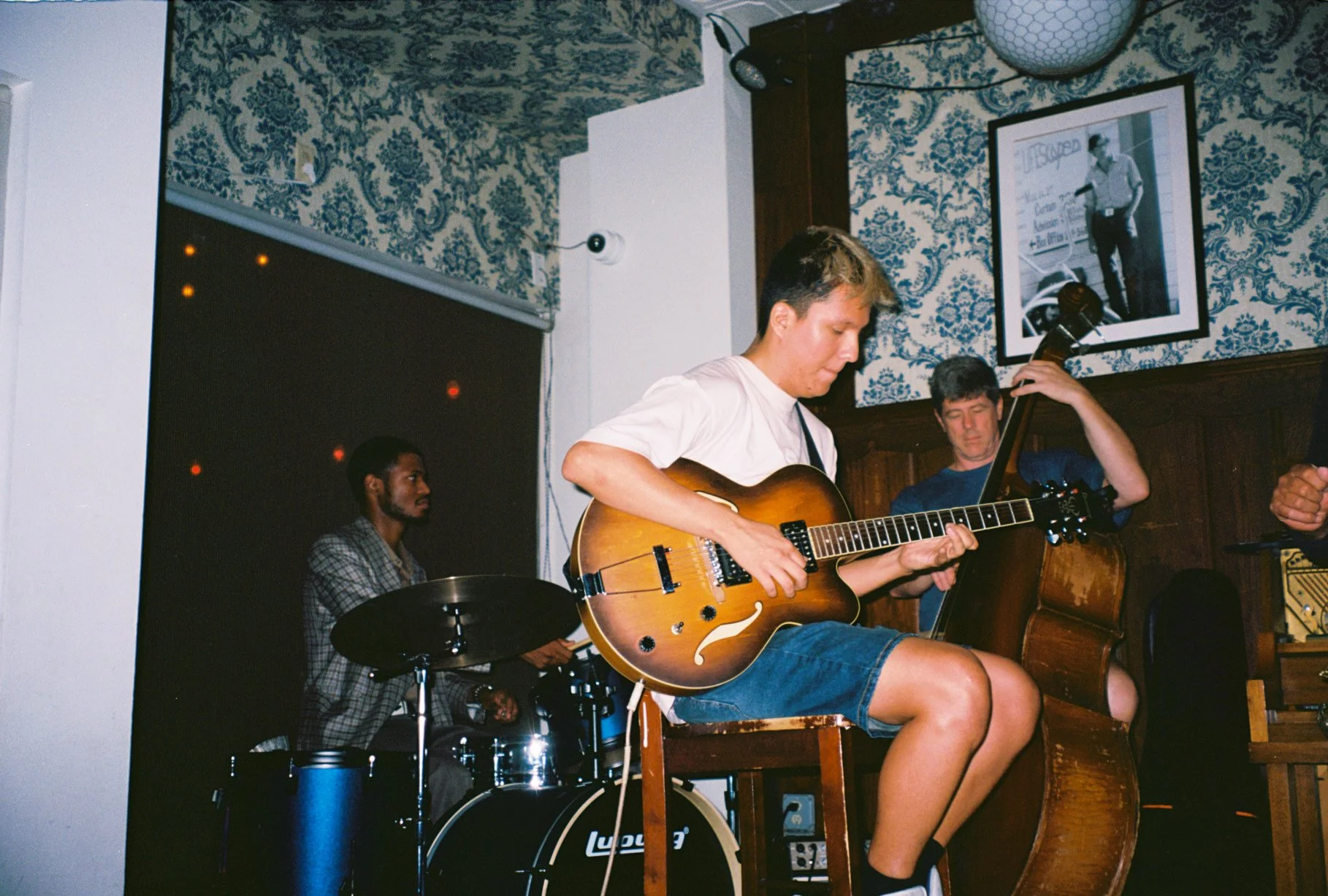 Three musicians performing live in a room with blue and white wallpaper; one plays drums, another plays a guitar, and the third plays a double bass.