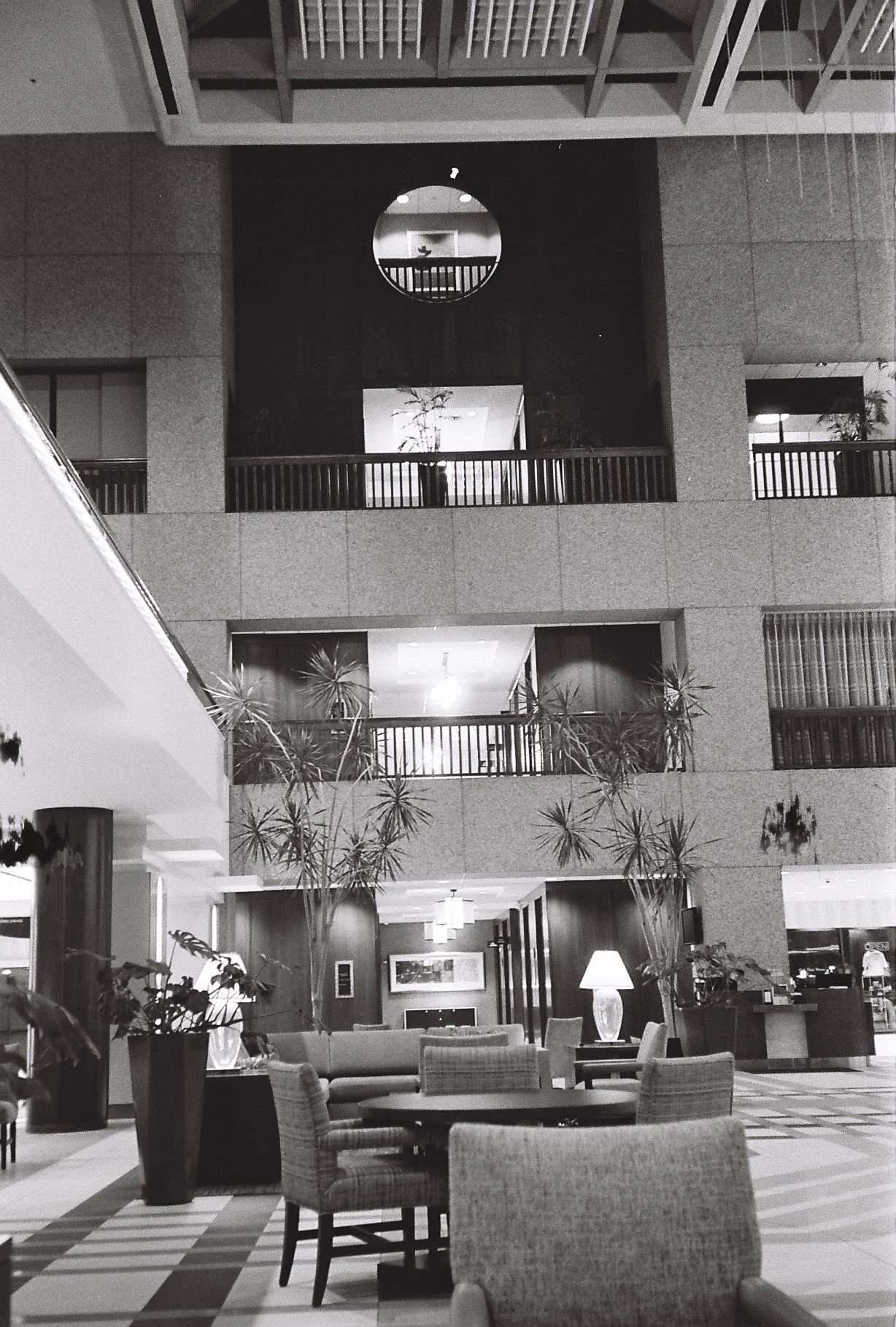 The interior of a hotel lobby with seating areas, potted plants, and a multi-level structure with balconies and railings, illuminated by lamps and ceiling lights.