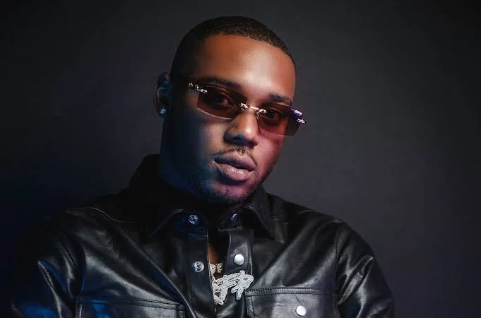 A man with short hair wearing sunglasses and a black leather jacket against a dark background.