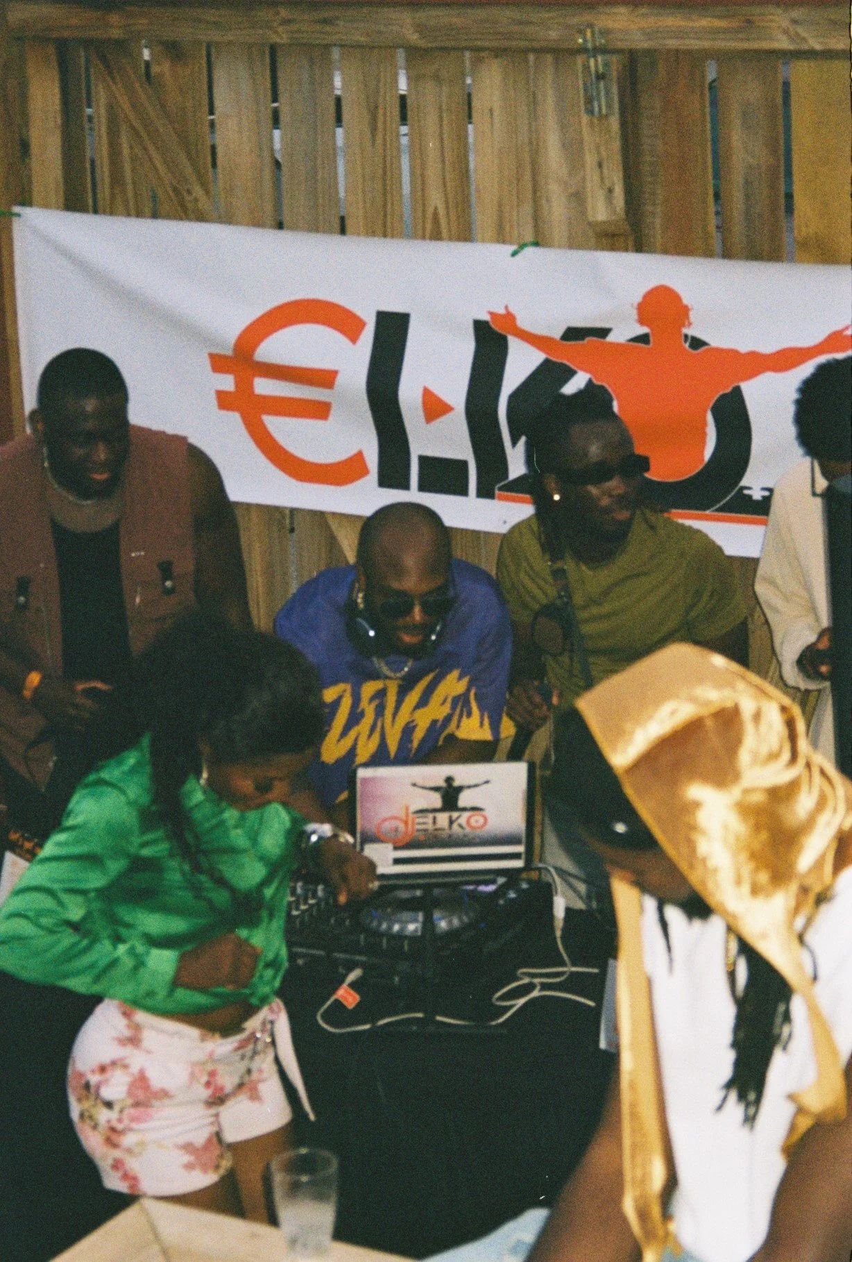 People at a party or event with a DJ booth, a banner with a euro symbol and a silhouette of a woman, and individuals in casual attire, some wearing sunglasses and headphones, holding food and drinks.