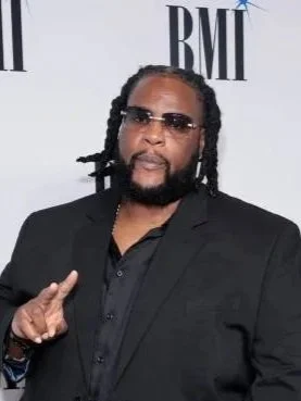 Man with dreadlocks and beard wearing sunglasses and a black suit posing at the BMI awards.