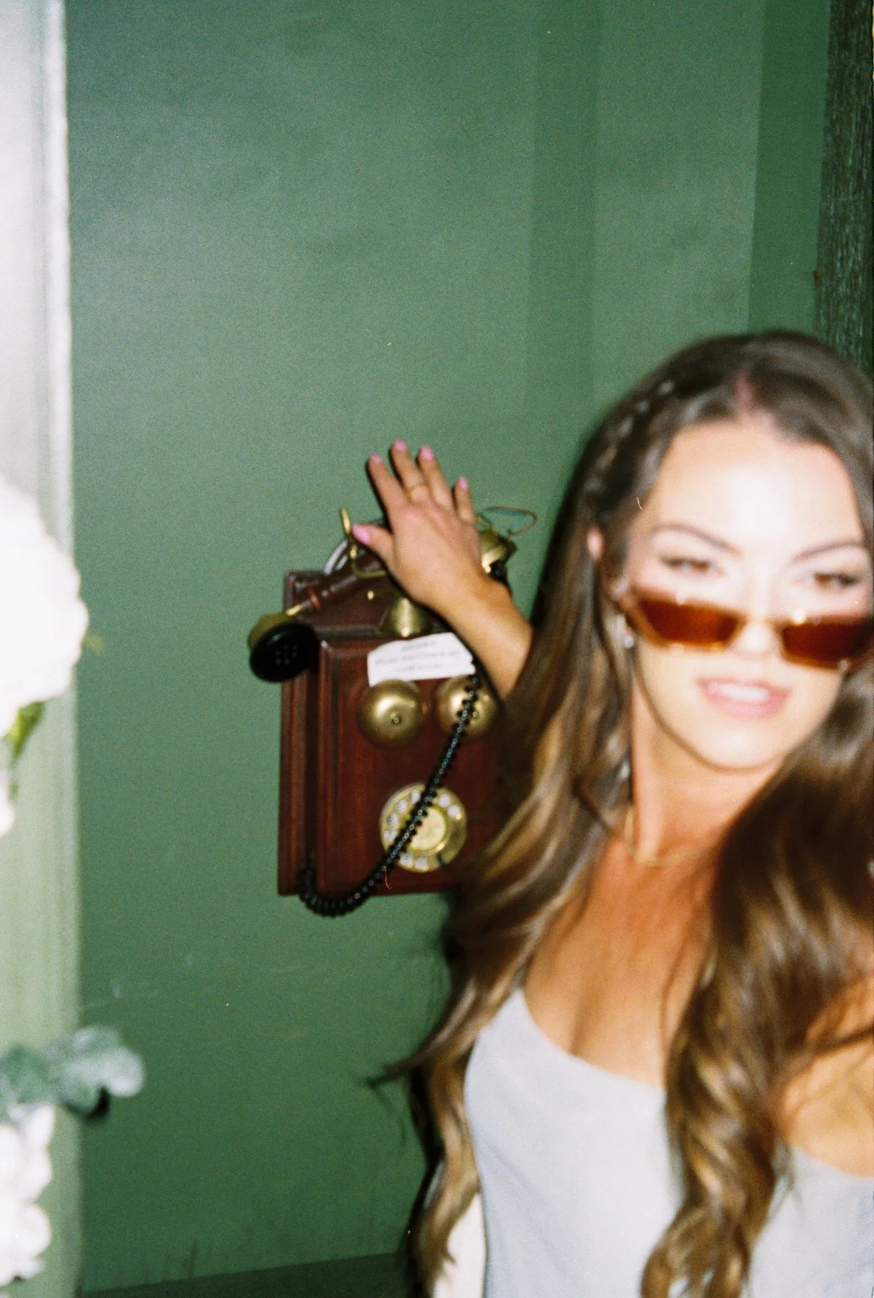 A woman with long brown hair wearing sunglasses, smiling, with her hand resting on an antique wall phone in a room with green walls.