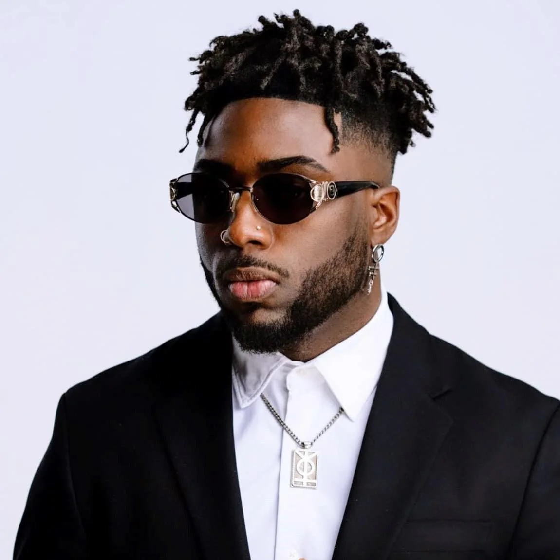 A young man wearing black sunglasses, a white shirt, a black blazer, and silver jewelry, including a chain and earrings, against a plain light background.