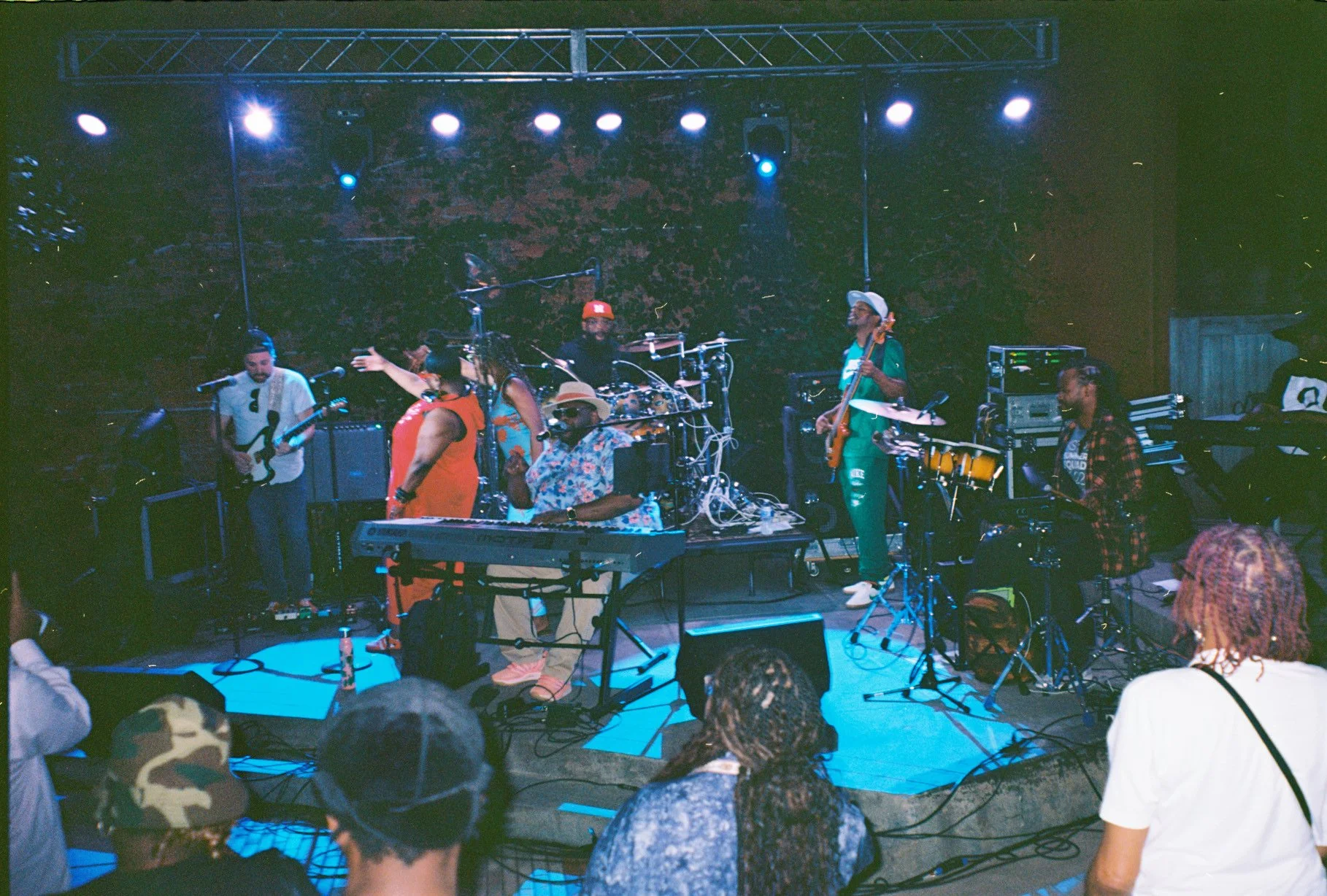A live band performs on stage in a dimly lit venue with several audience members watching. The band includes musicians playing keyboard, electric guitar, drums, bass, and percussion. The stage has various musical equipment and lighting, creating a vi
