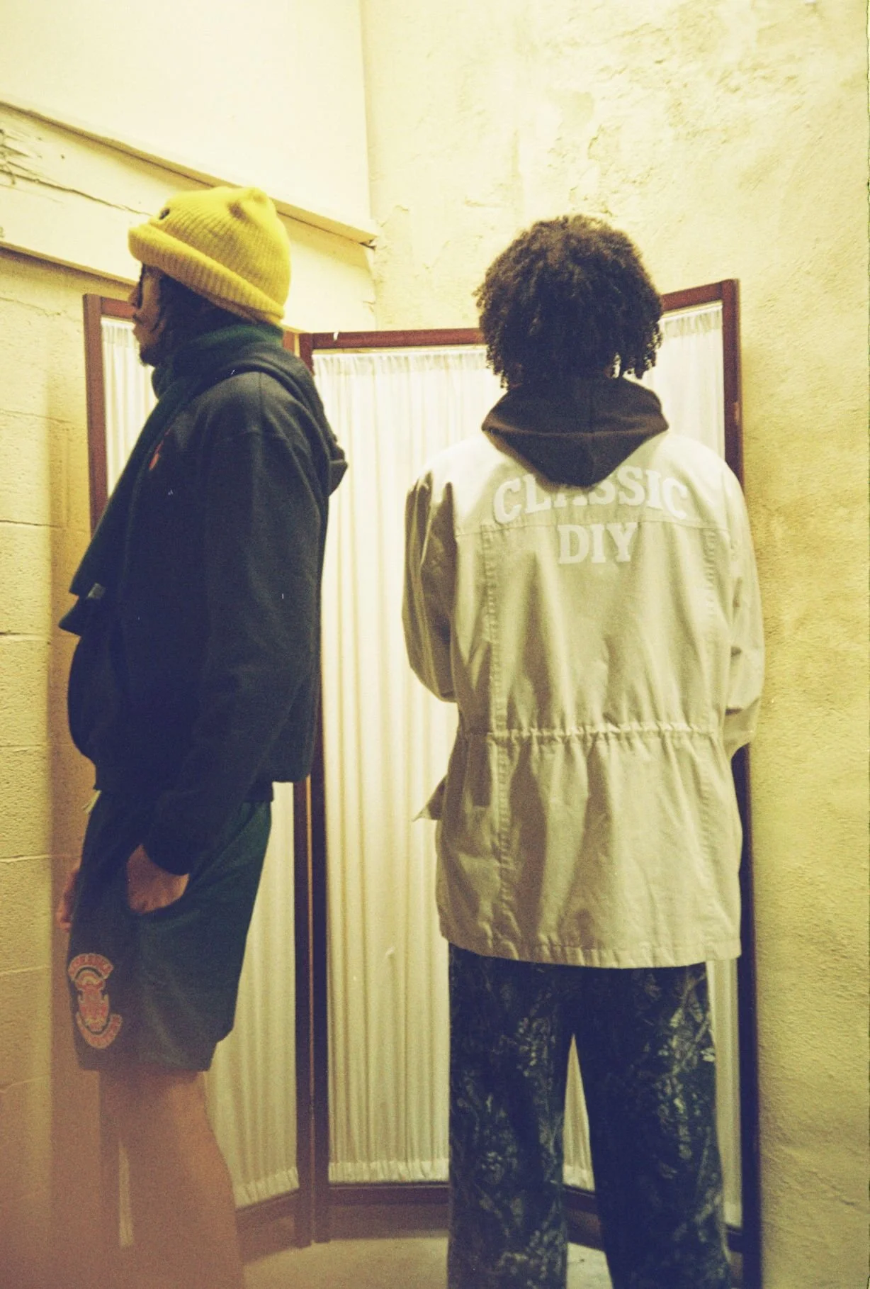 Two individuals standing in front of a mirror with beige curtains, one wearing a yellow beanie and dark jacket, the other wearing a beige jacket with 'CLASSIC DIY' printed on the back and patterned pants.