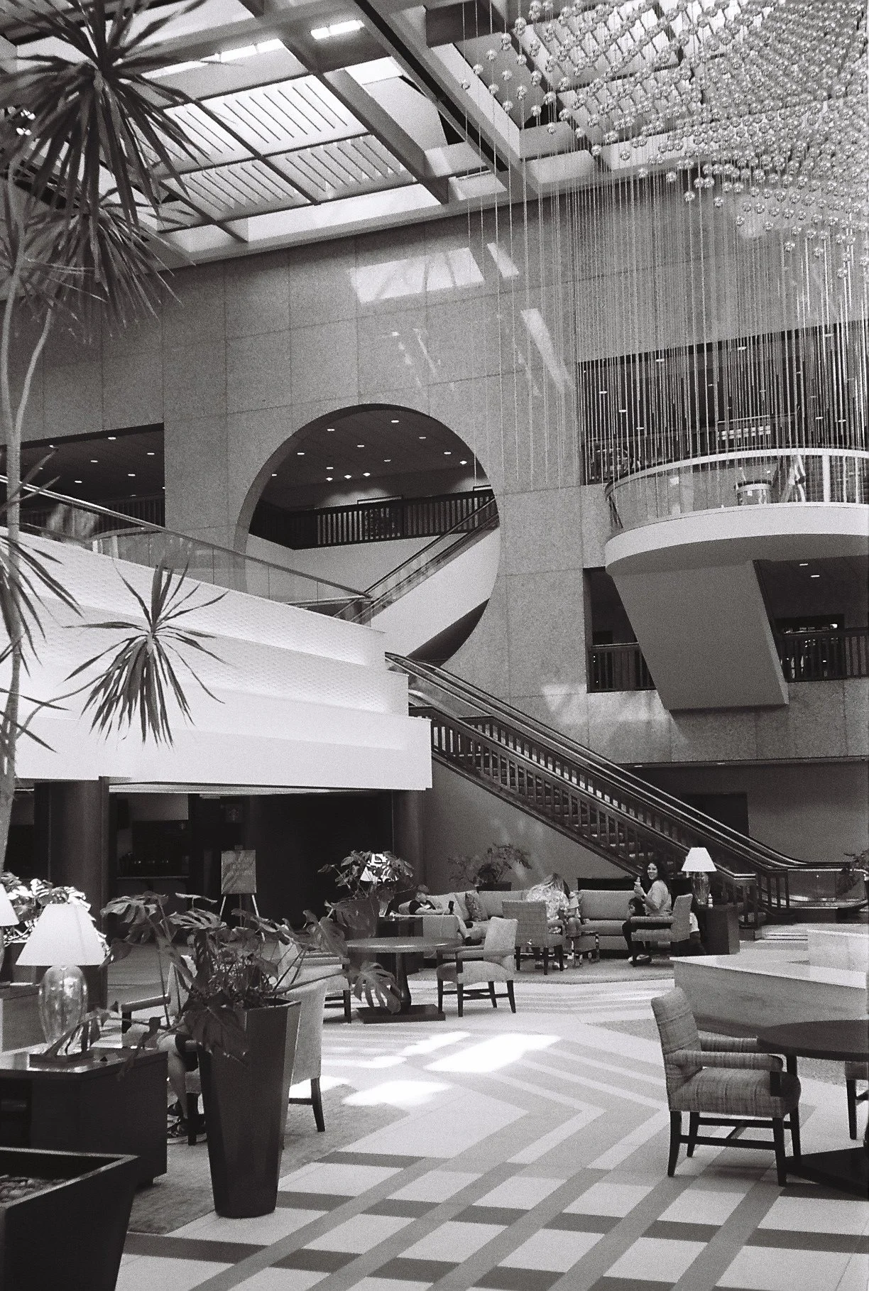 Interior of a hotel lobby with seating areas, potted plants, a staircase, and a large modern chandelier hanging from the ceiling.