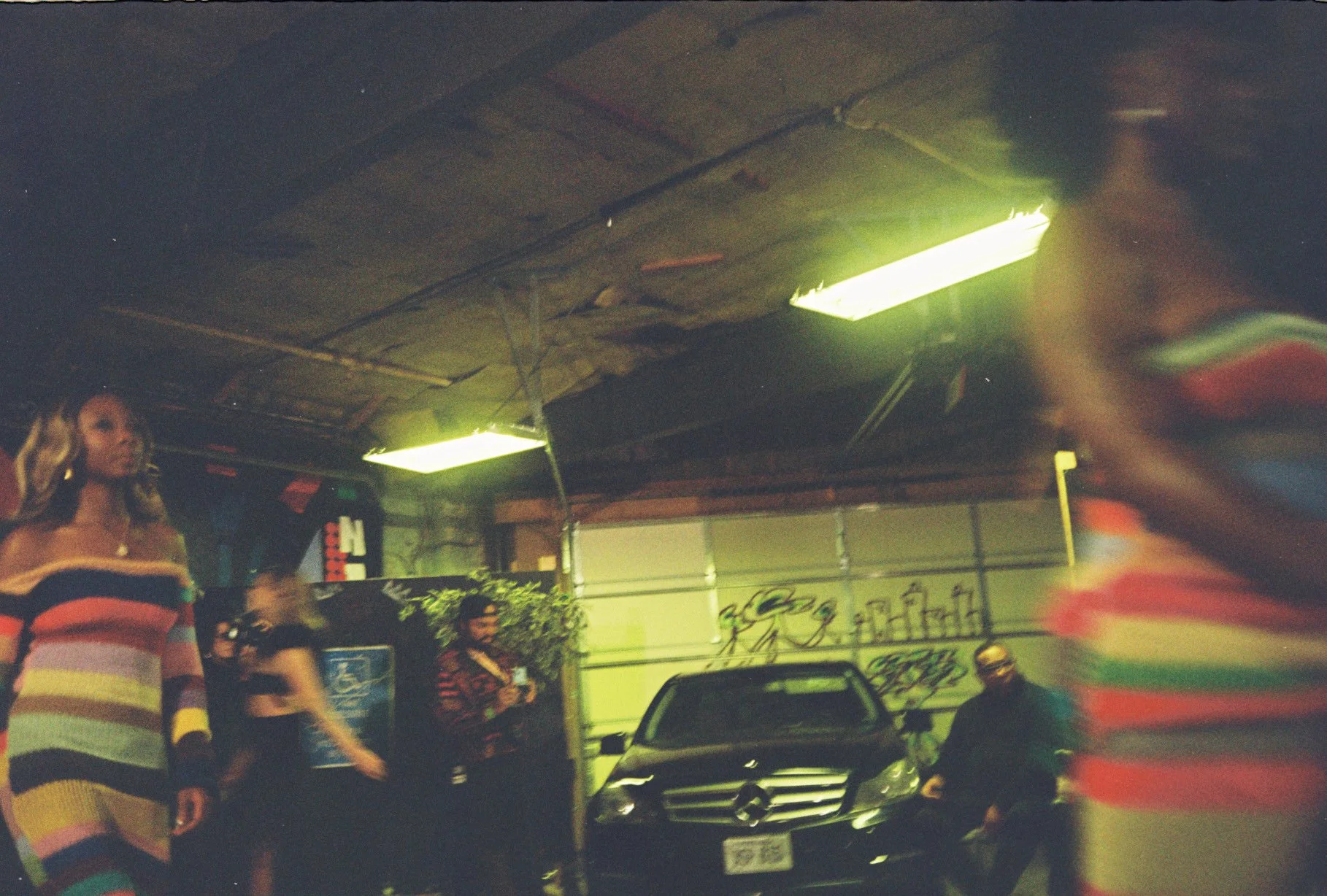 People inside a garage with a black Mercedes-Benz SUV, fluorescent lights on the ceiling, and graffiti on the wall in the background.