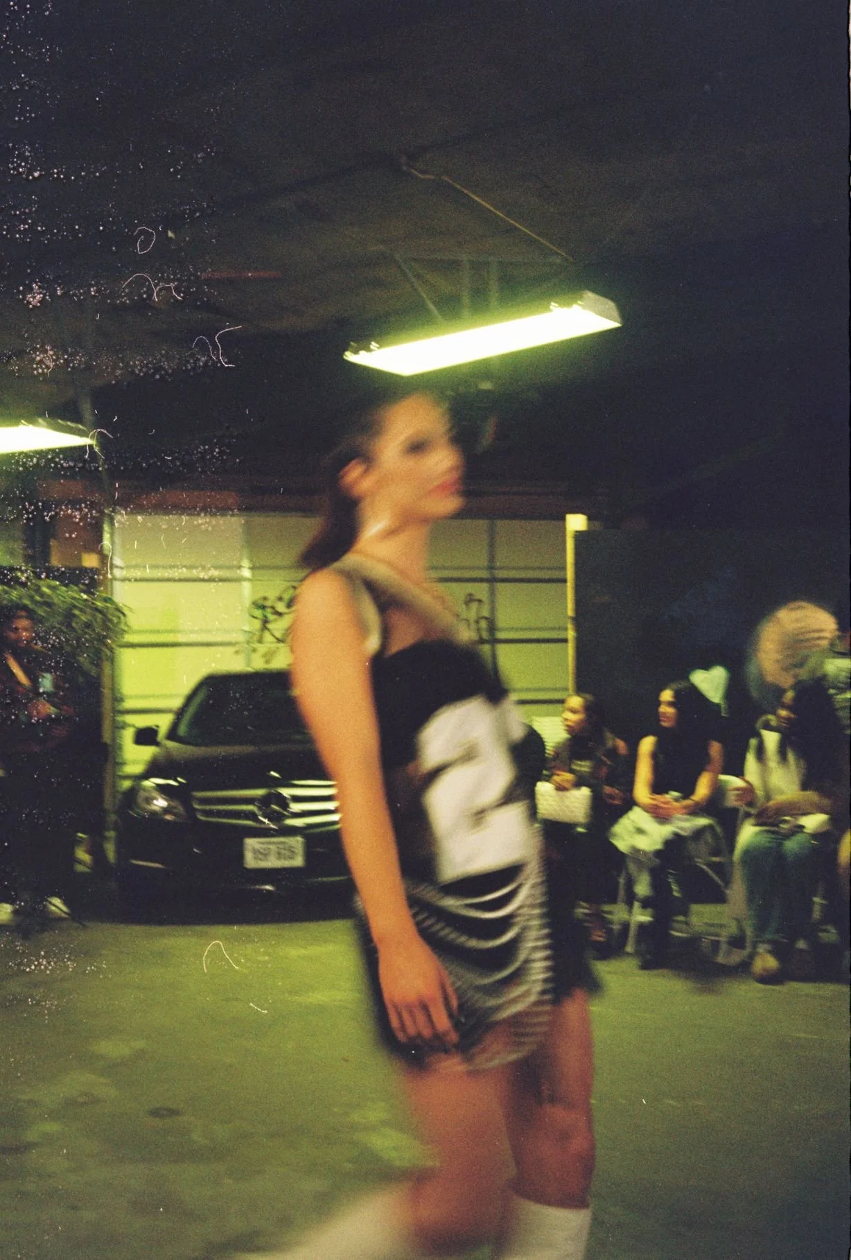 A woman in a black and white striped dress standing in a dimly lit garage with several people sitting on chairs, a black Mercedes-Benz car, and a fluorescent ceiling light. Raindrops are visible on a glass surface in the foreground.