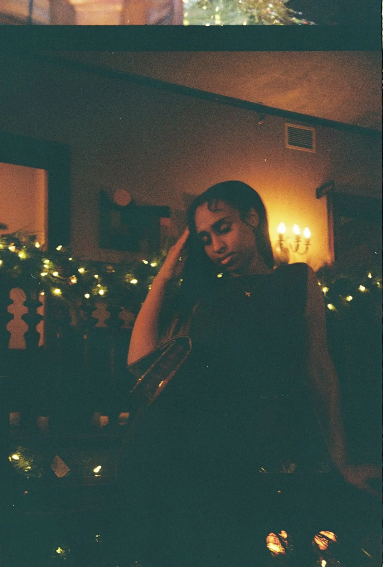 A woman sitting by a window at night, with warm lighting and Christmas lights on a railing behind her, looking downwards with her hand in her hair.