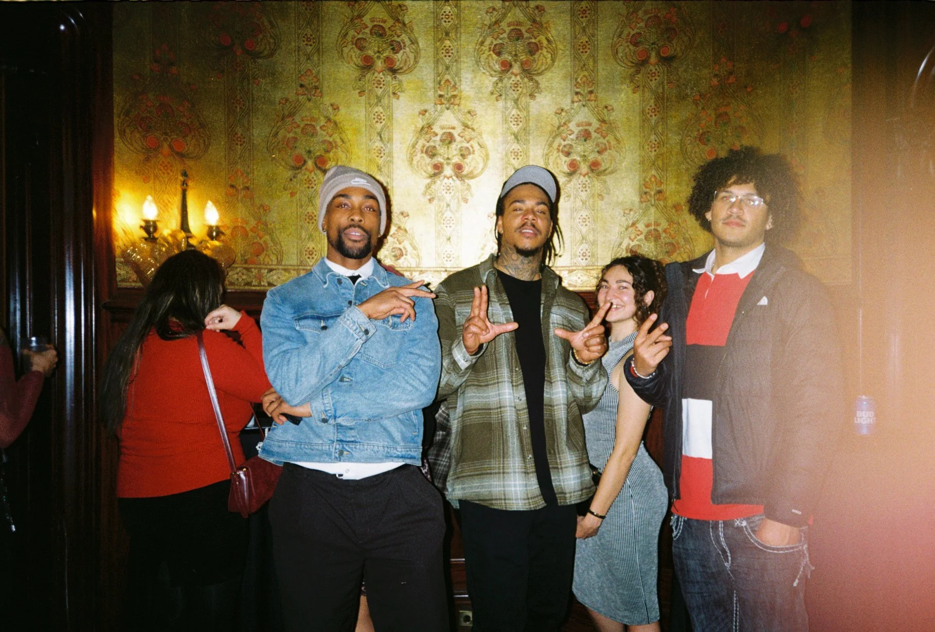 Four young adults posing together at a social gathering, standing in front of ornate wallpaper and warm lighting. The group includes two men and two women, all appearing relaxed and happy.