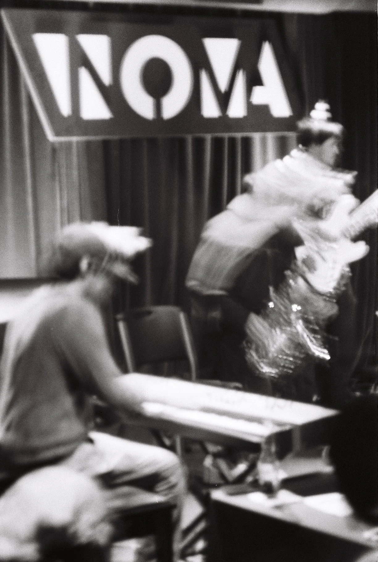 A black and white photo showing two women dressed as princesses, both wearing tiaras and shiny skirts, sitting at a table at an event. There is a large sign with the word 'NOMA' hanging behind them, and an audience is visible in the foreground.