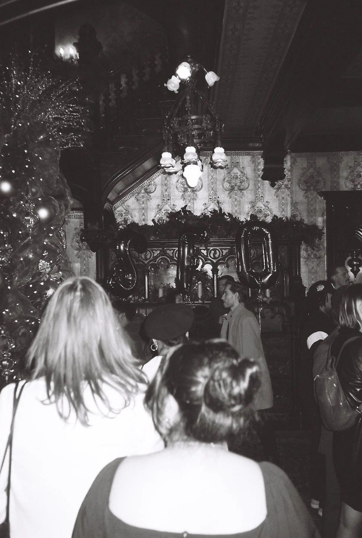 People gathered in a decorated, ornate room with a large mirror, a chandelier, and balloons, likely celebrating a special occasion.