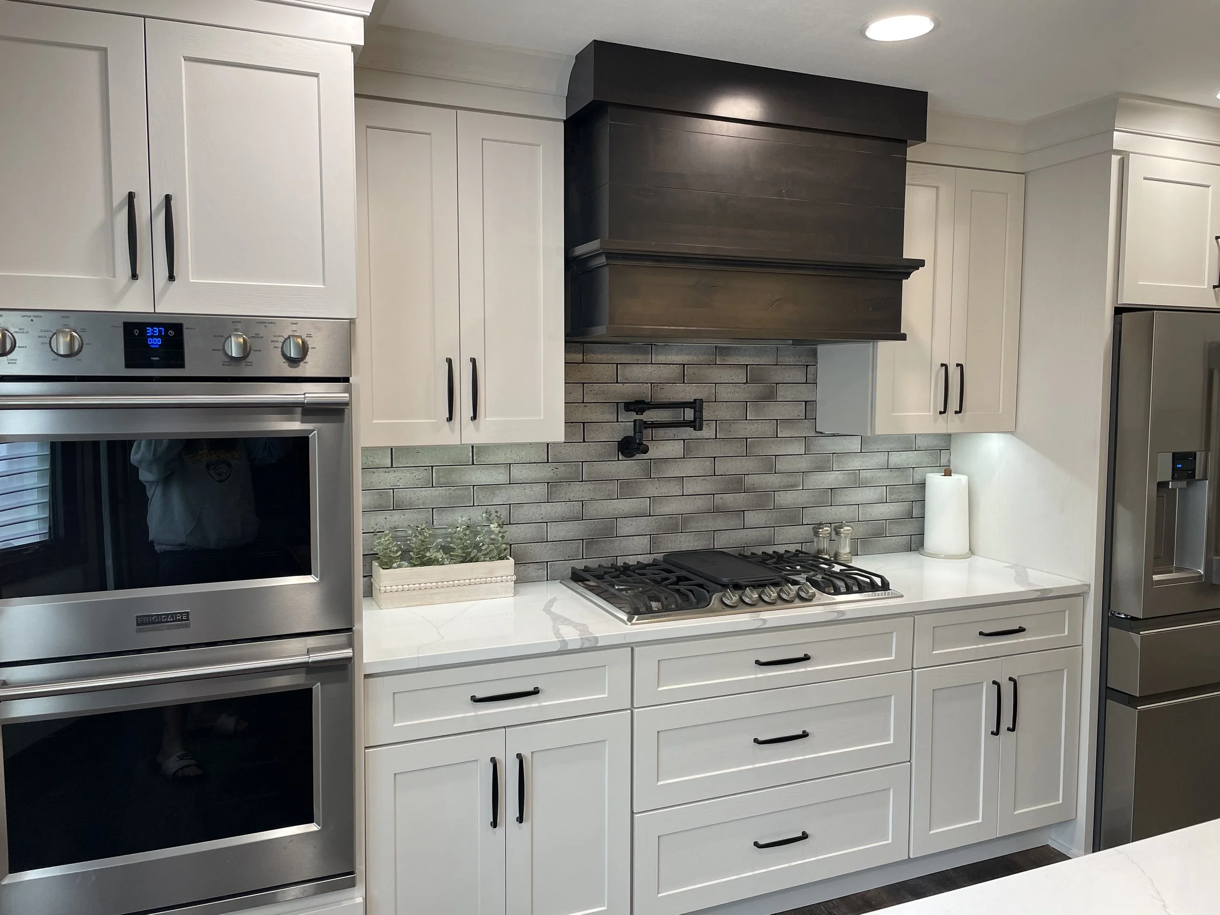 Kitchen with white cabinets, black handles, a stainless steel oven, gray brick backsplash, a black range hood, and a built-in stovetop, with a paper towel roll on the countertop.