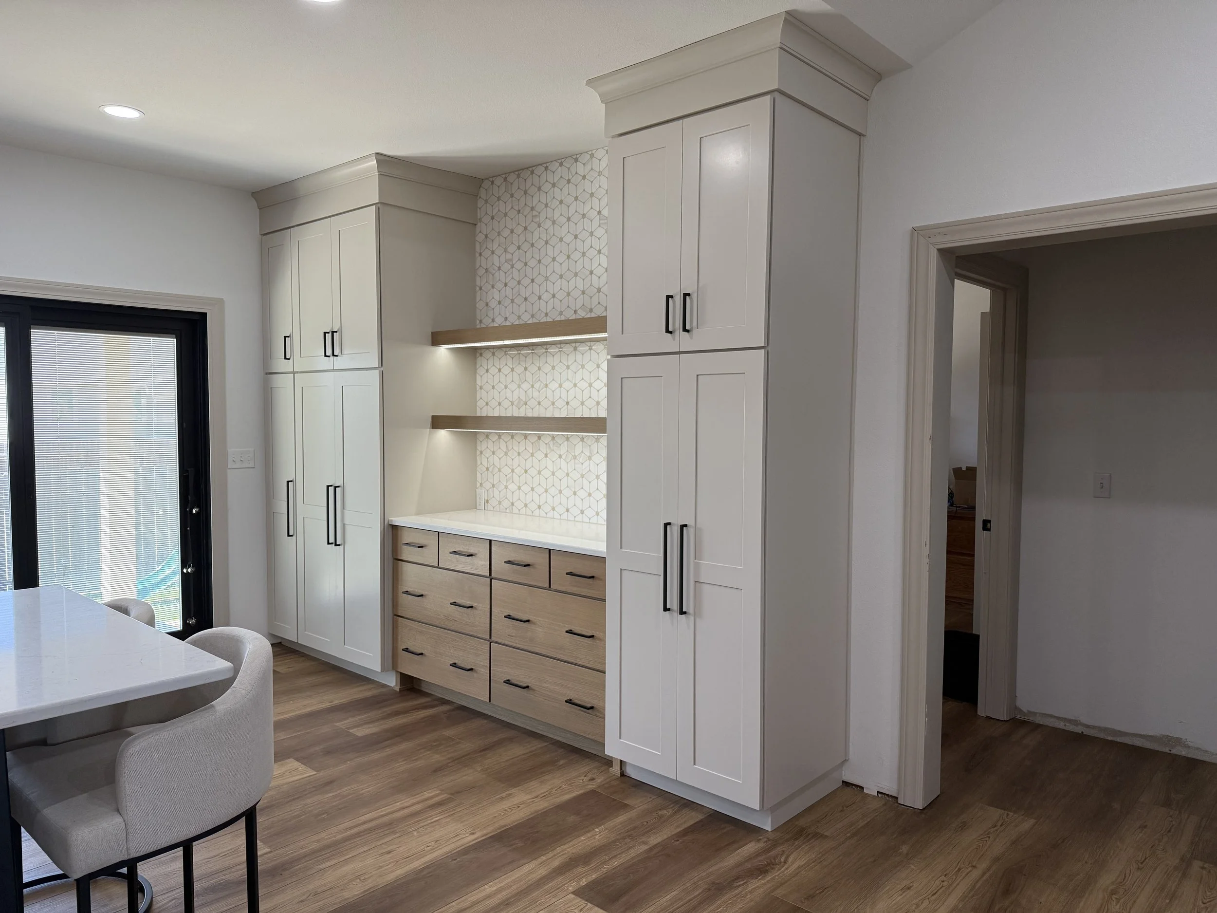 Modern kitchen with white cabinets, a decorative tile backsplash, and a wooden dresser-style cabinet. A door with blinds and a corner of a dining table with chairs are visible.
