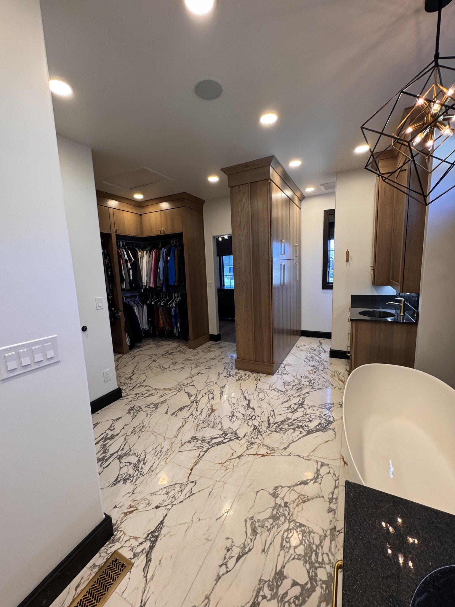 Interior of a modern bath and walk-in closet with marble flooring, wooden cabinets, a small black countertop with a sink, and a hanging geometric chandelier.