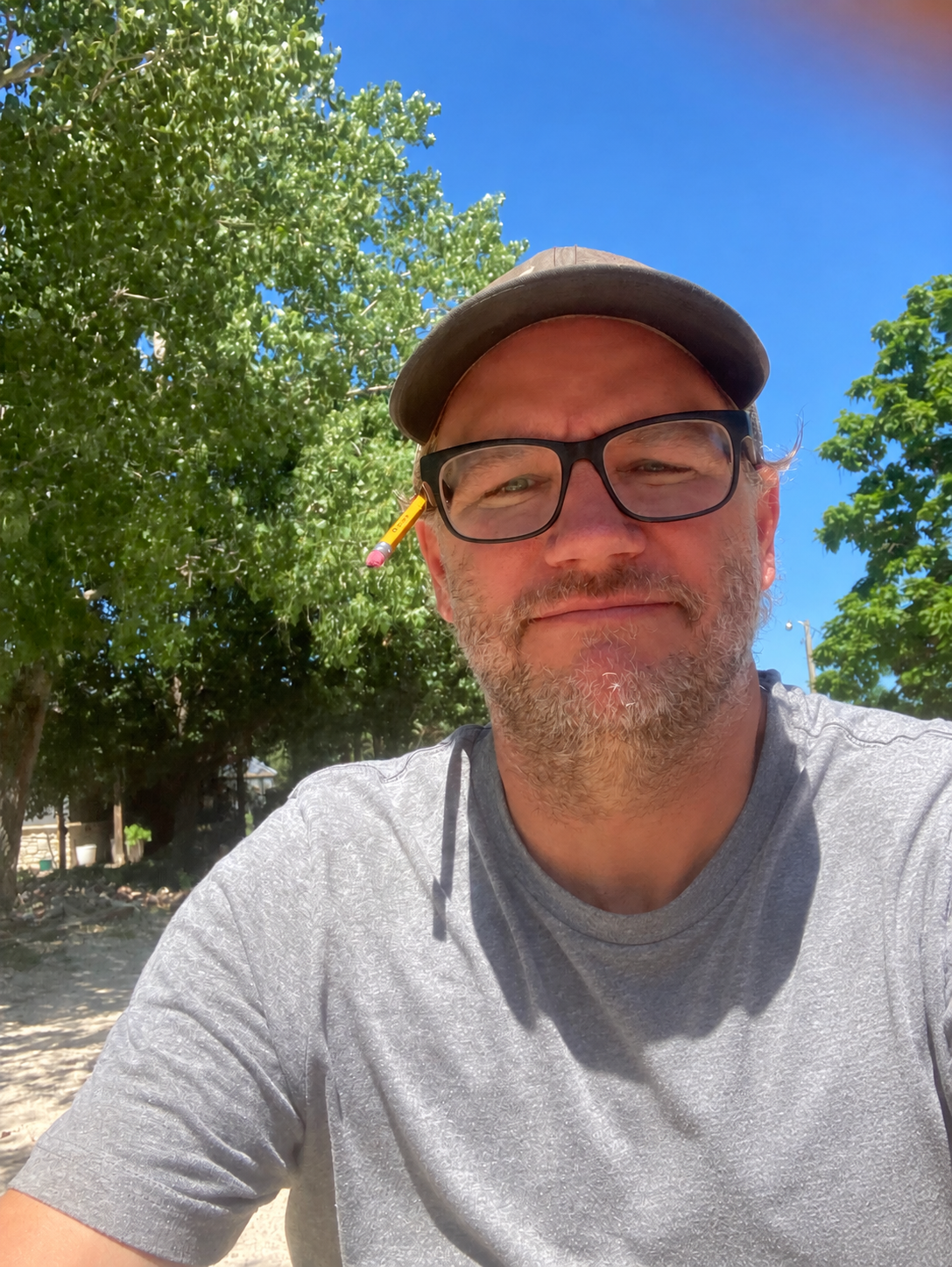 A middle-aged man with glasses, a baseball cap, and a pencil tucked behind his ear is taking a selfie outdoors on a sunny day. There are tall green trees and a clear blue sky in the background.