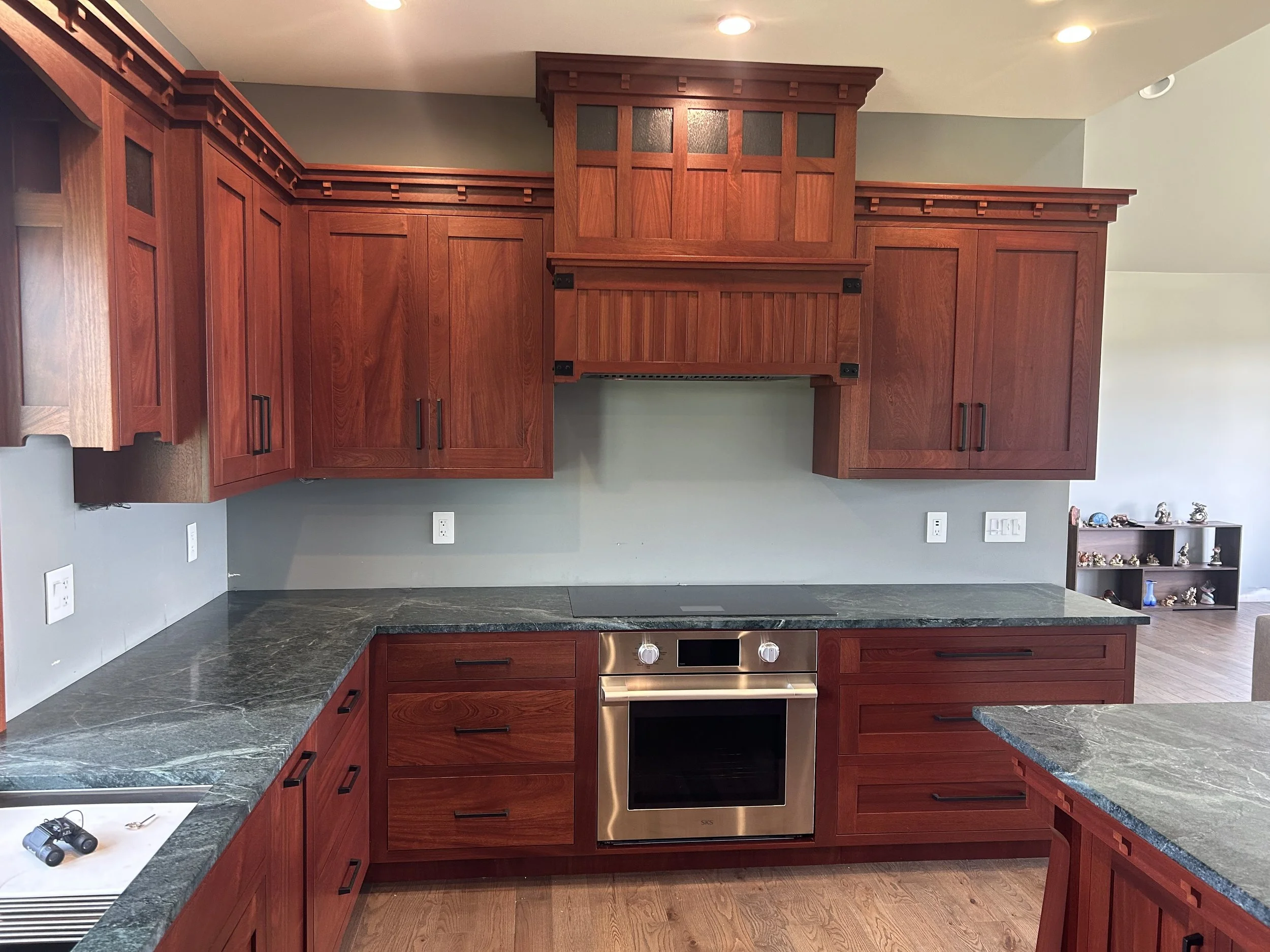 Kitchen with wooden cabinets, black countertops, stainless steel oven, and a small black bookshelf with decorative items