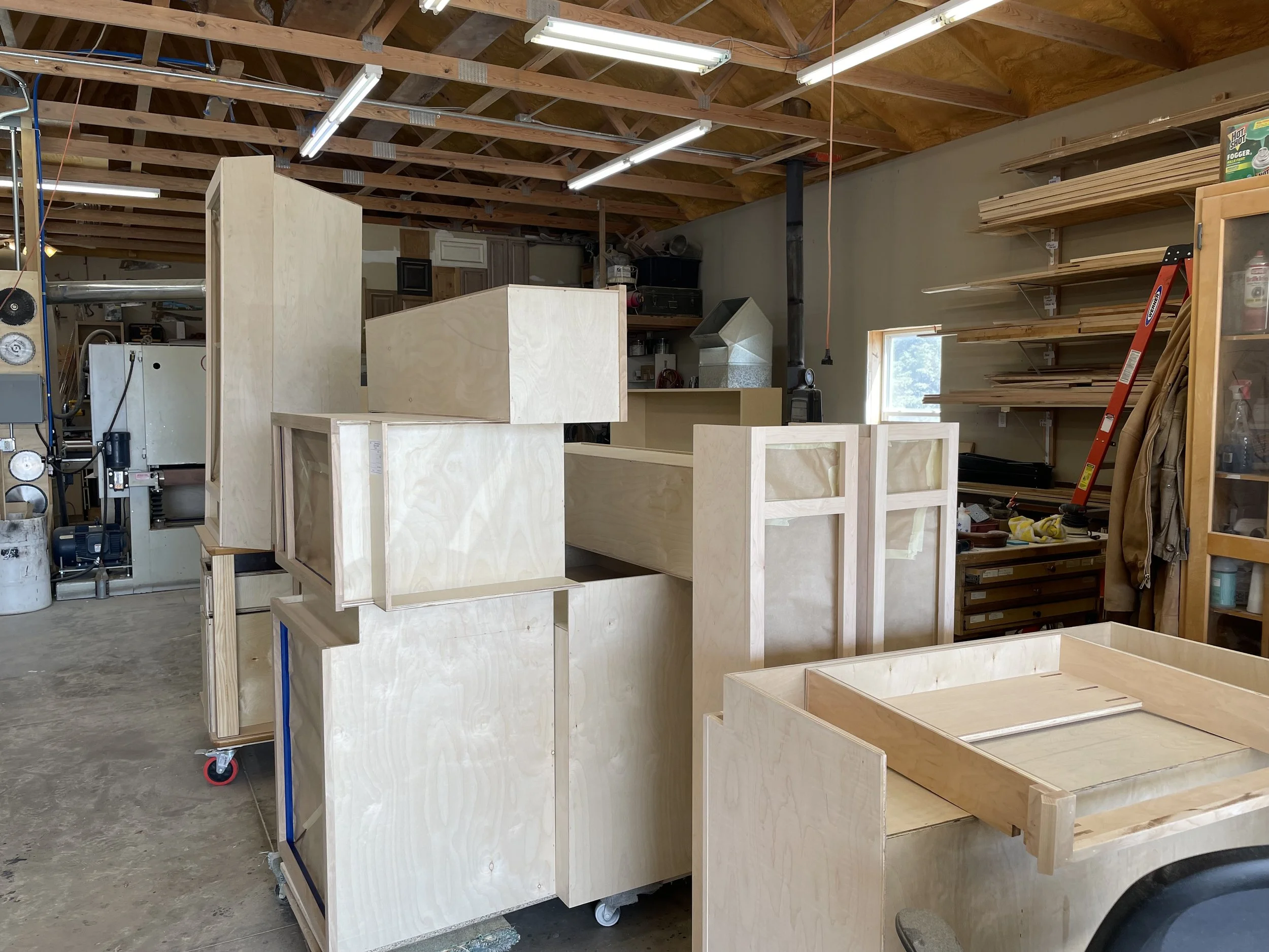 Interior of a woodworking shop with unfinished wooden frames and furniture, shelves with tools and supplies, and a ladder.
