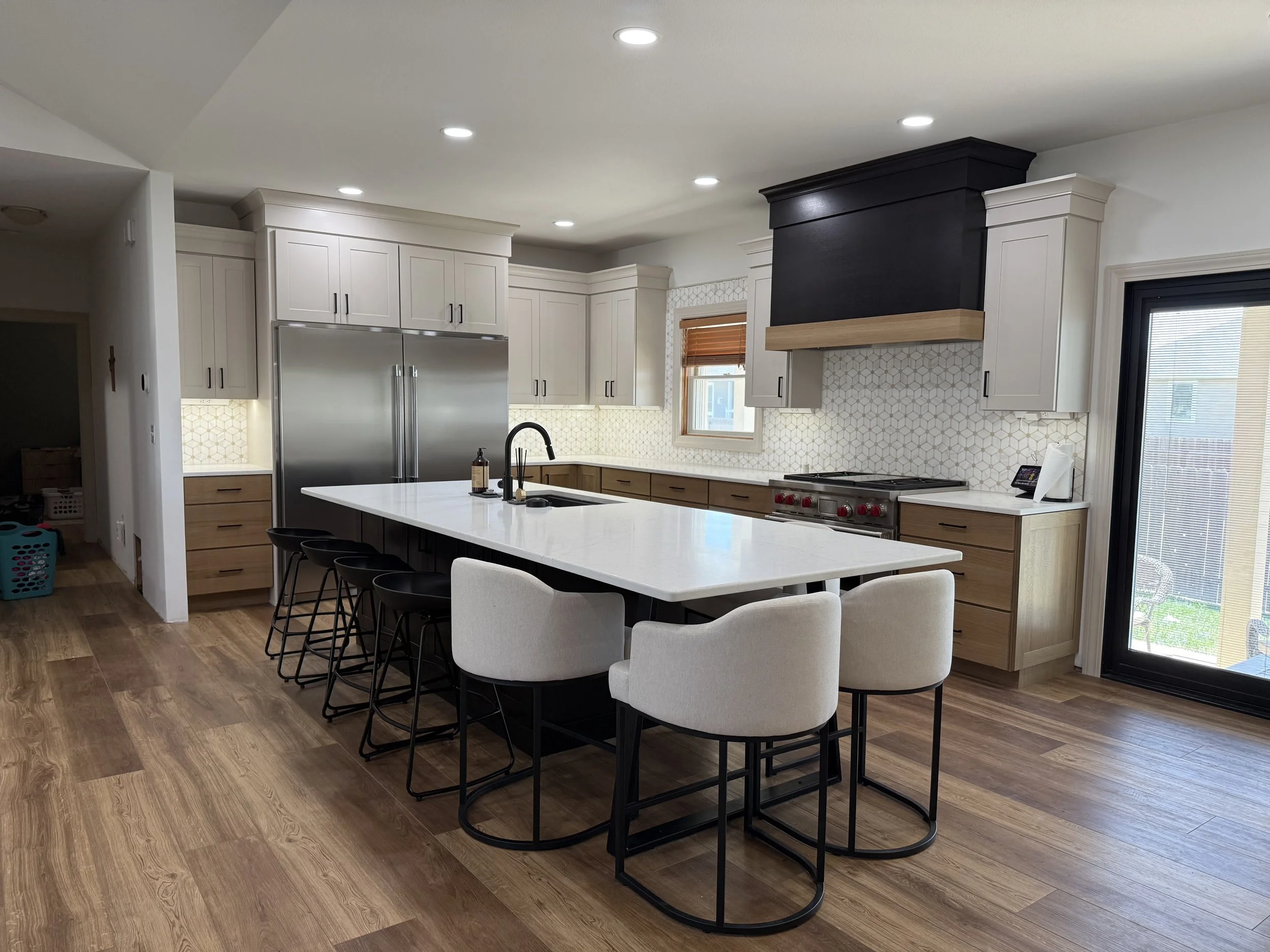 Kitchen with white cabinets, black accents, and a large white island with seating, stainless steel appliances, wood flooring, and a black chimney hood over the stove.