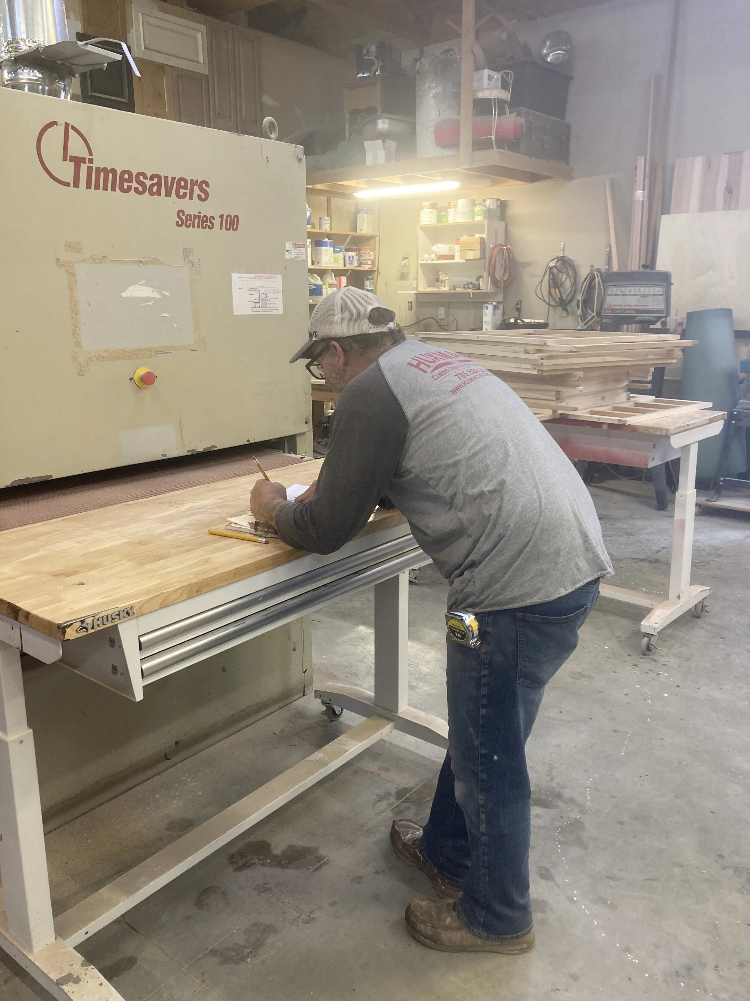 Jeremy in a workshop wearing a gray and black shirt, jeans, a cap, and glasses, measuring and marking a piece of wood on a workbench, with woodworking tools and supplies in the background.